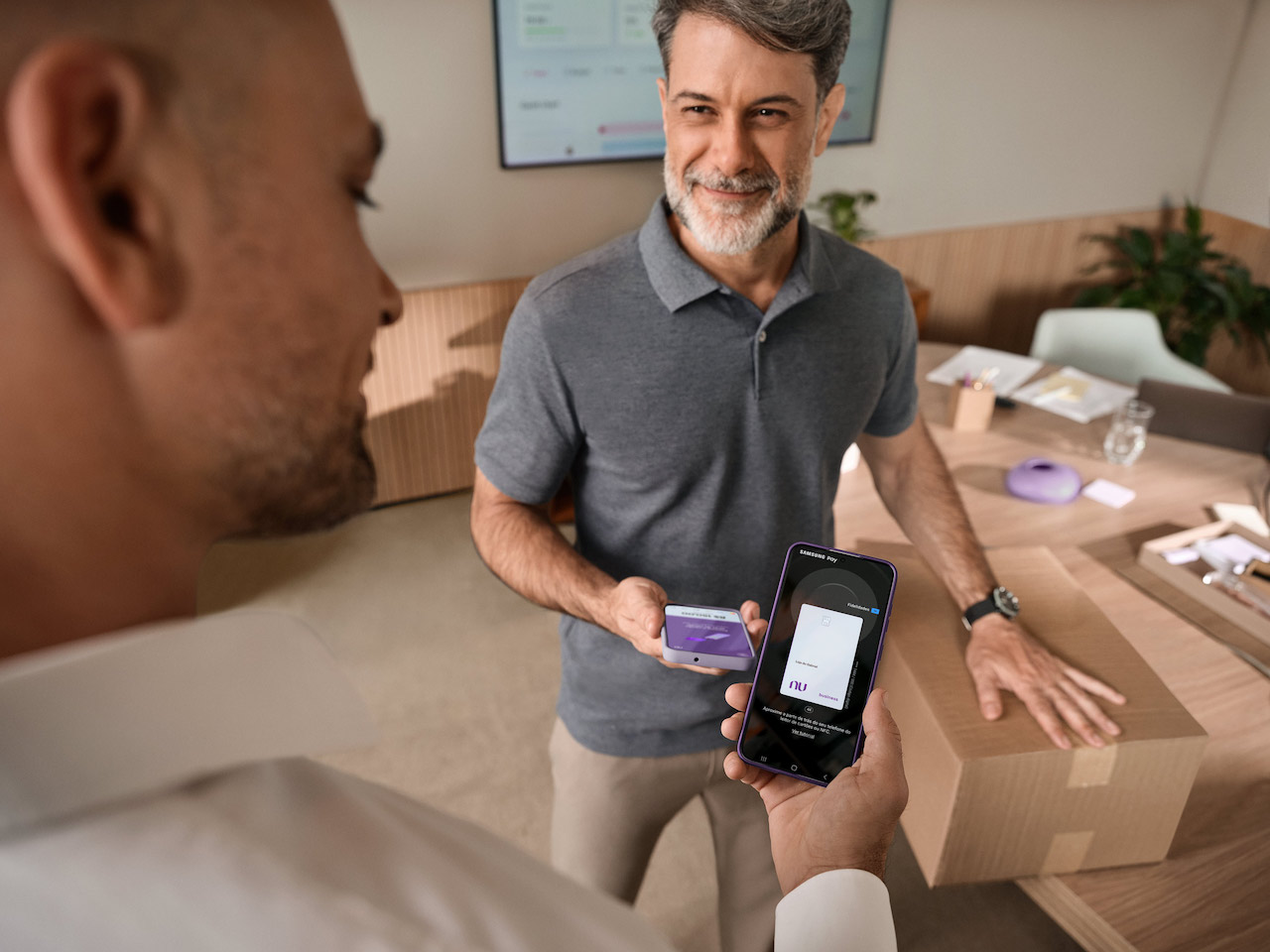 Em primeiro plano, imagem mostra um homem de costas vestindo uma camisa branca segurando um celular. Ele olha para outro homem, que está de frente, sorrindo, e segurando um celular em uma das mãos. Ele veste uma camisa polo cinza e calça caqui.