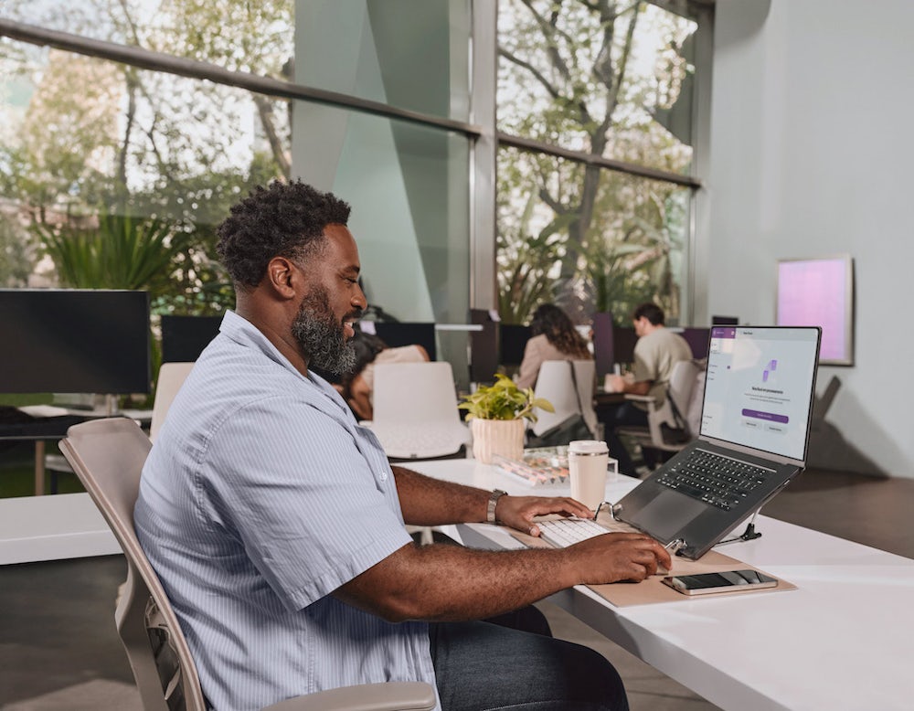 Em primeiro plano, homem negro sentado à mesa de um escritório usando um notebook. Ao fundo, outras pessoas trabalham em mesas do escritório. O homem veste uma camisa azul claro e calças pretas. Ele tem barba e cabelos curtos pretos.