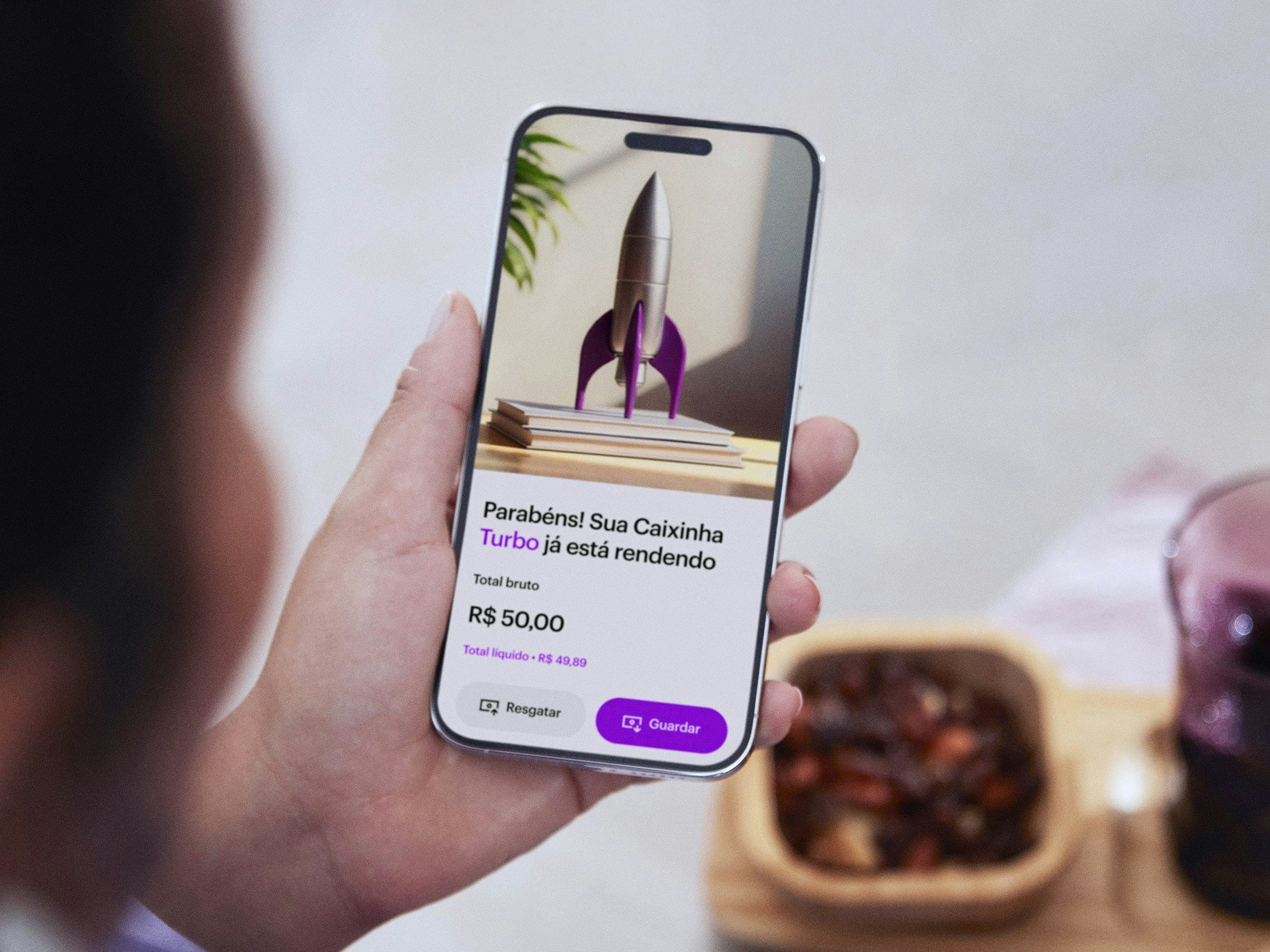 Mão branca segura um celular em primeiro plano. A tela do celular mostra a seção do app do Nubank sobre Caixinha Turbo. Nessa tela, há uma foto de foguete.