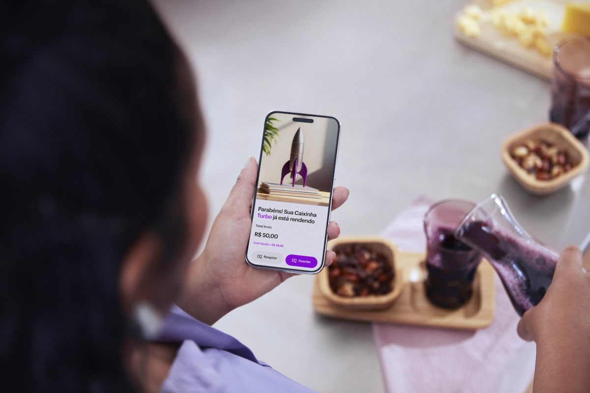 Mulher negra de costas segura um celular com o app do Nubank aberto na seção de Caixinha Turbo. Com a outra mão, ela coloca um suco de uva no copo. O copo está em uma mesa ao lado de potinhos de madeira com frutas.