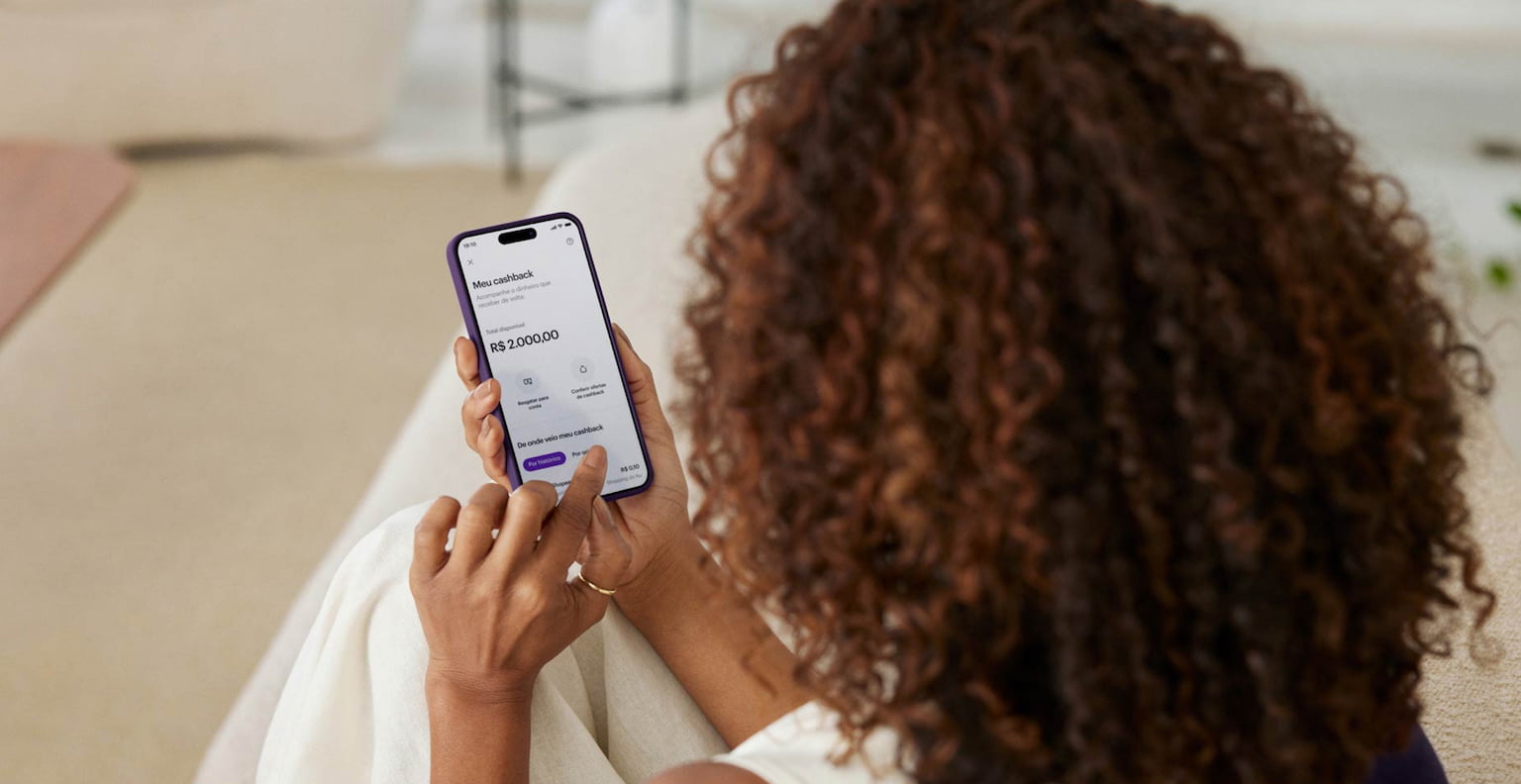 Mulher de costas sentada em um sofá segurando o celular, que está em evidência. Na tela do celular, o App do Nu está aberto na seção de Cashback do Cartão de Crédito Ultravioleta.