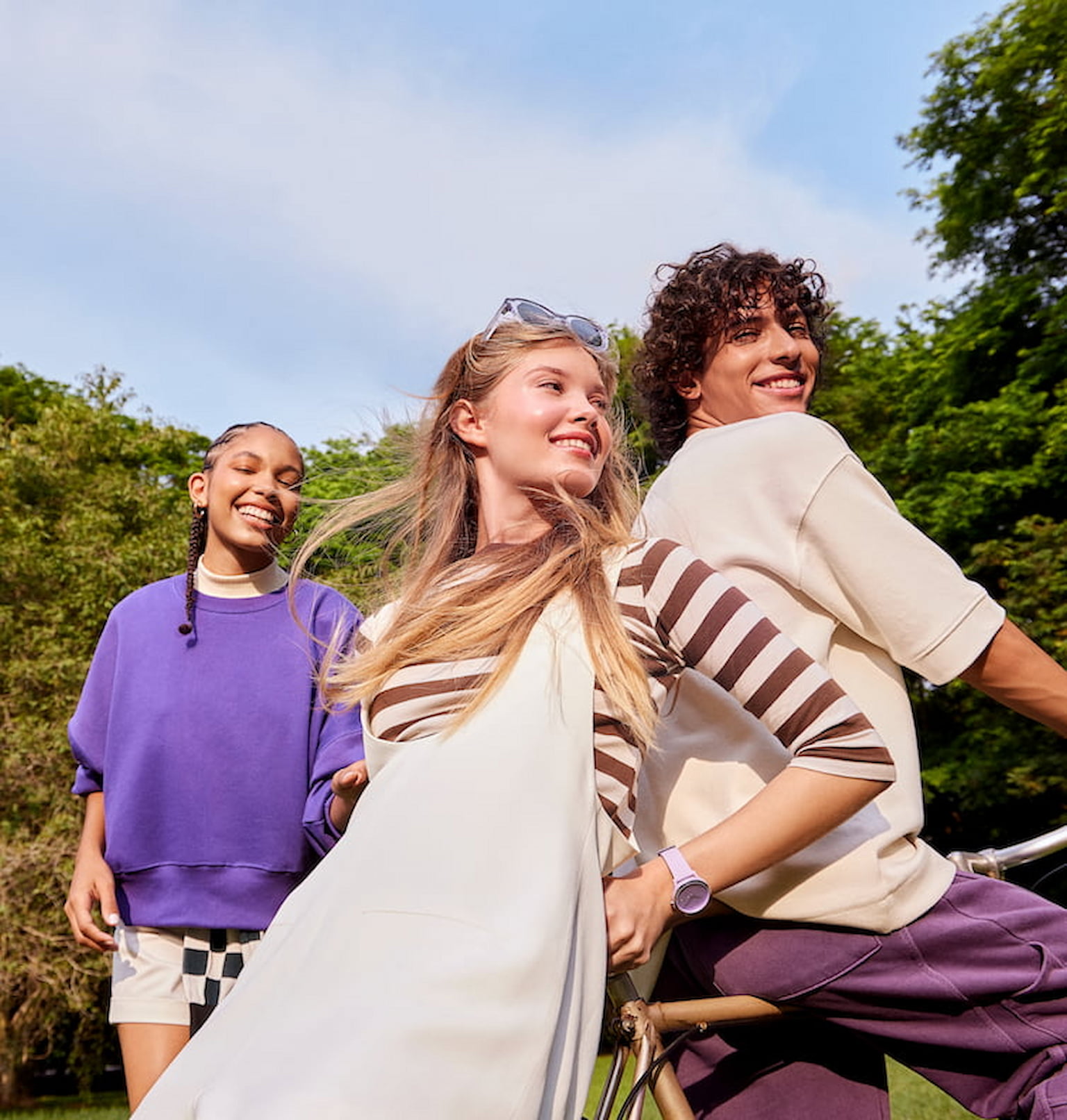 Duas meninas e um menino sorrindo em um parque. Uma das meninas está vestindo uma blusa roxa e a outra está com uma blusa bege com listras marrons horizontais com um vestido bege por cima. O menino está de costas, em cima de uma bicicleta, vestindo uma camiseta bege e uma calça roxa.
