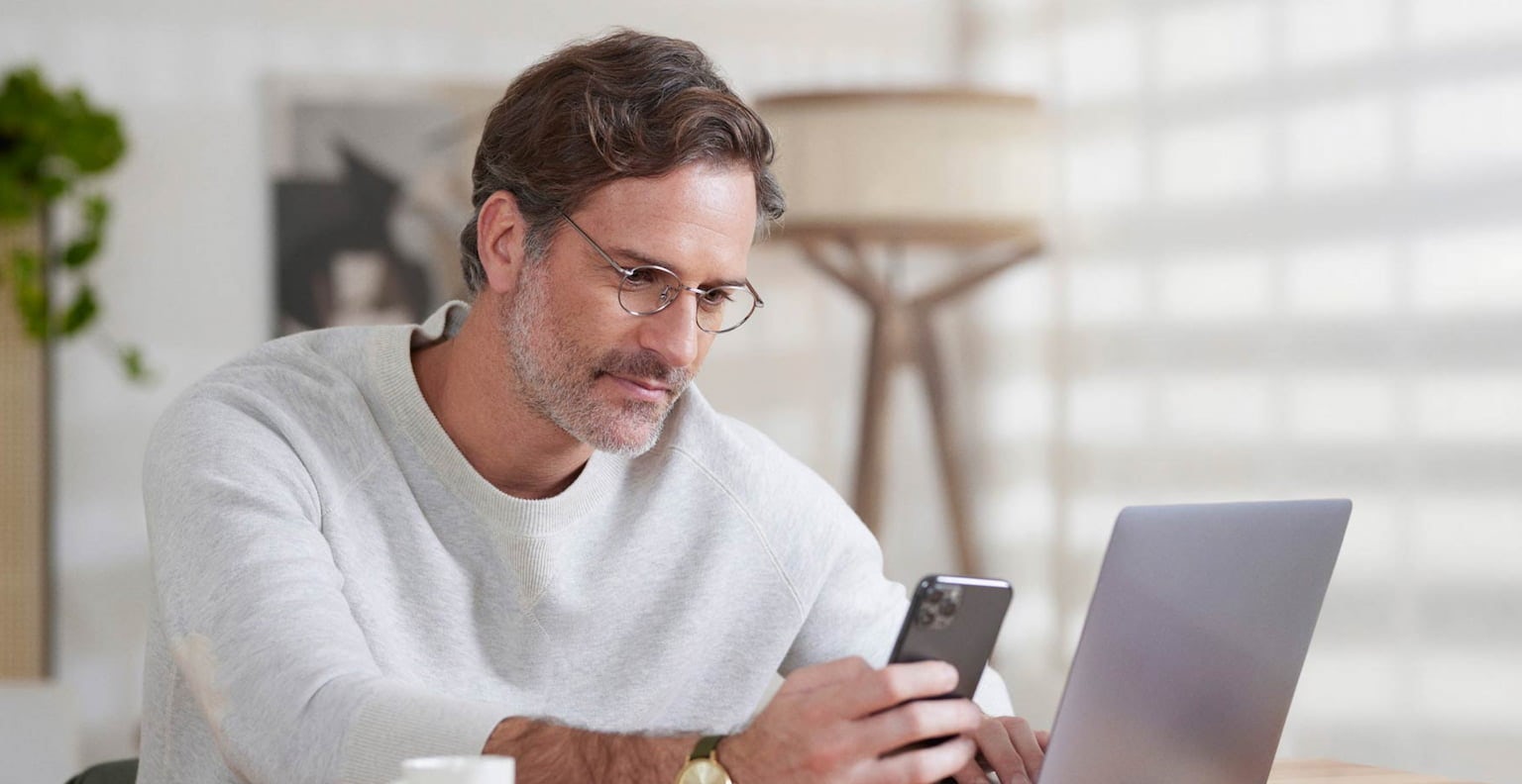 Homem de óculos sentado em um escritório olhando para a tela do celular que está em uma de suas mãos. A outra mão está em cima do teclado do notebook. Na mesa, uma xícara de café, uma agenda e uma caneta.
