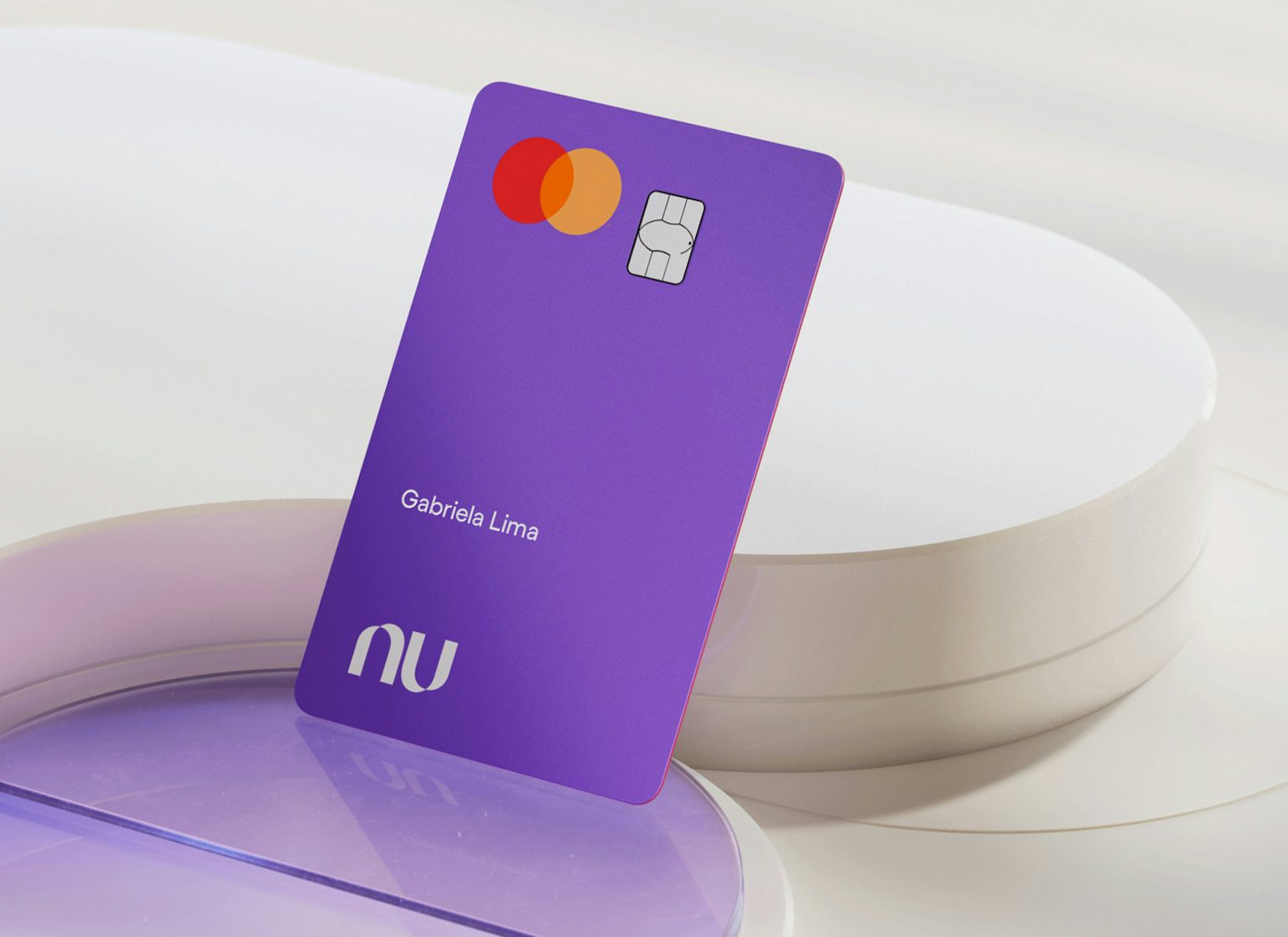 Cartão do Nubank visto de cima posto sob superfícies geométricas circulares. O cartão está com a face principal virada para cima, com o logo da bandeira Mastercard, chip e nome do cliente.