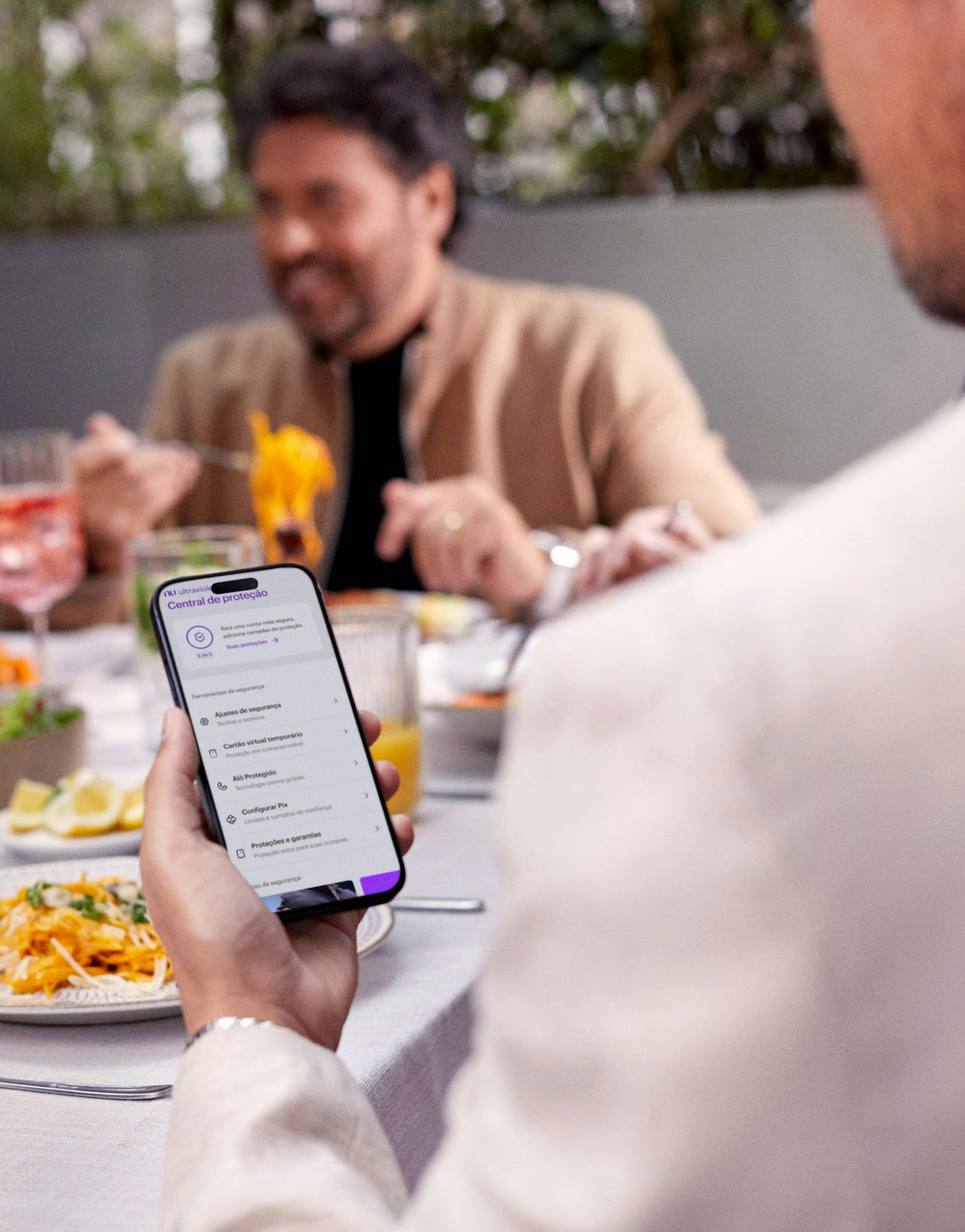 Homem de costas olha para o celular, que está em sua mão esquerda sobre uma mesa com drinks e comidas. Ele está checando a Central de Proteção Ultravioleta, no App do Nu. Ao fundo e de frente para ele, um homem rindo enquanto almoça entre amigos..