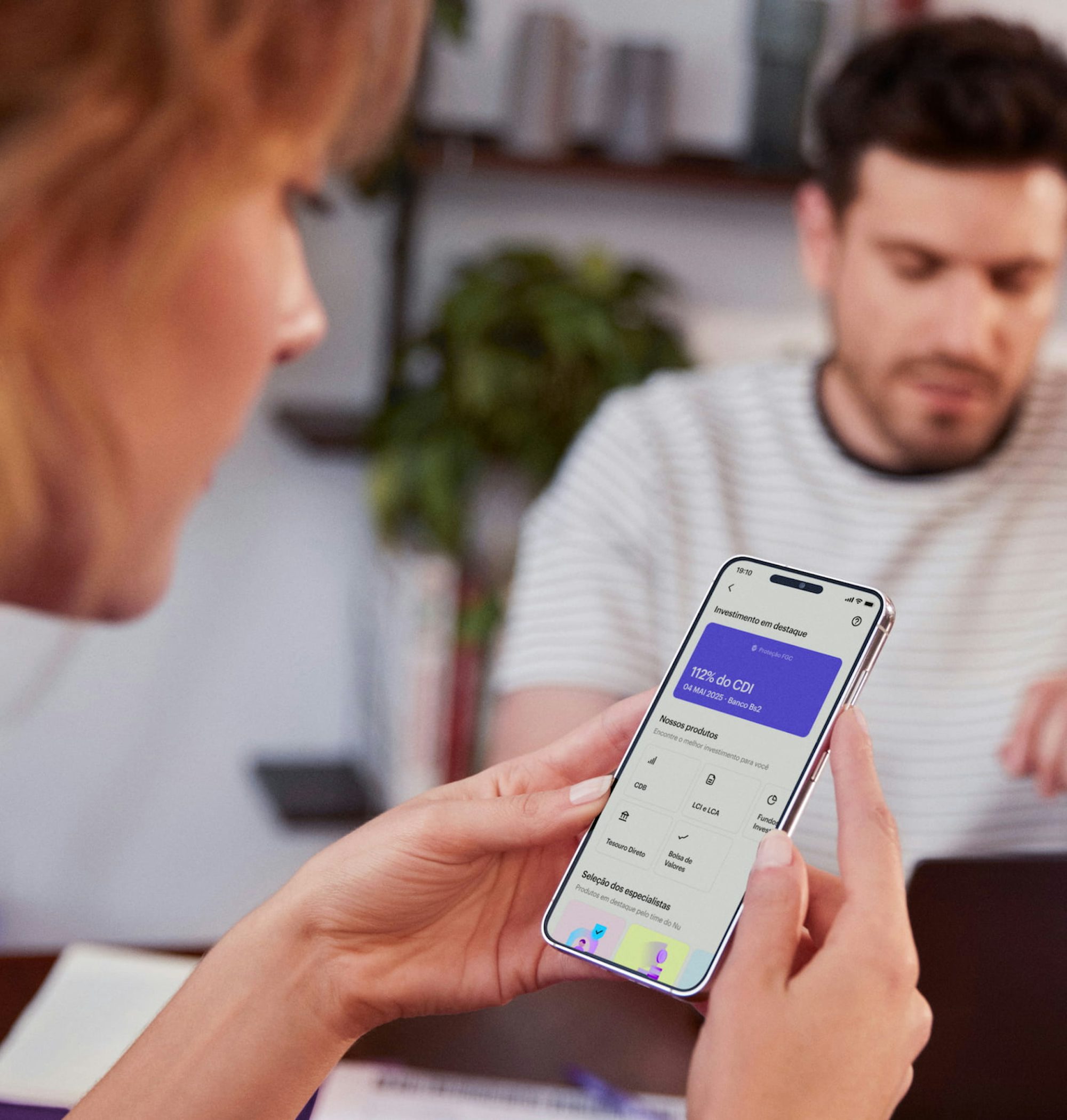 Perfil de uma mulher branca de cabelos curtos segura com as duas mãos um celular e visualiza sua conta no aplicativo do Nubank, ao fundo e desfocado um homem branco olha para um notebook em cima de uma mesa de madeira com um caderno aberto e alguns papéis espalhados.
