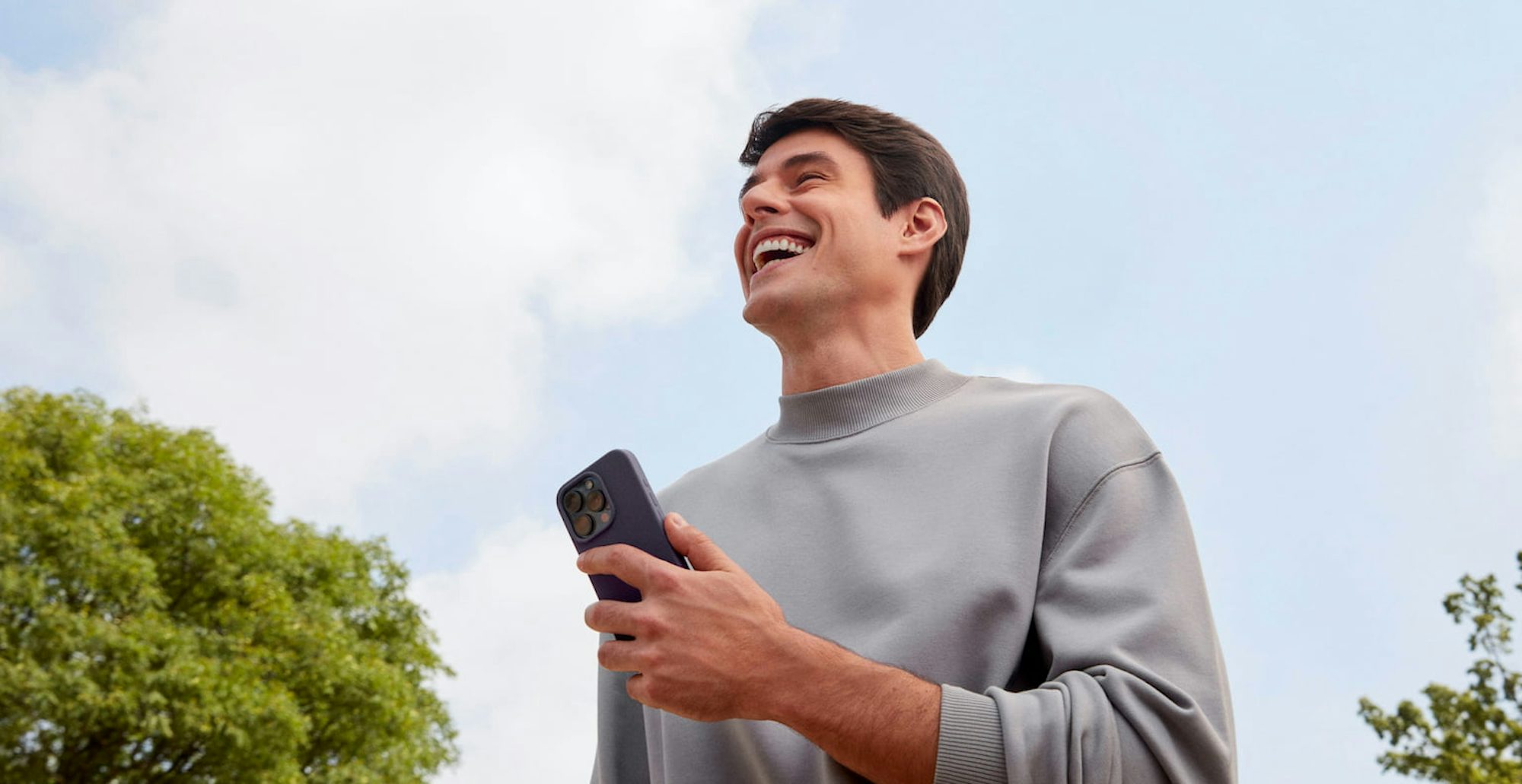 Homem branco sorrindo olhando para frente enquanto segura com a mão esquerda um celular, em um ambiente externo a luz do dia com nuvens no céu e uma árvore a sua esquerda e outra árvore a sua direita.