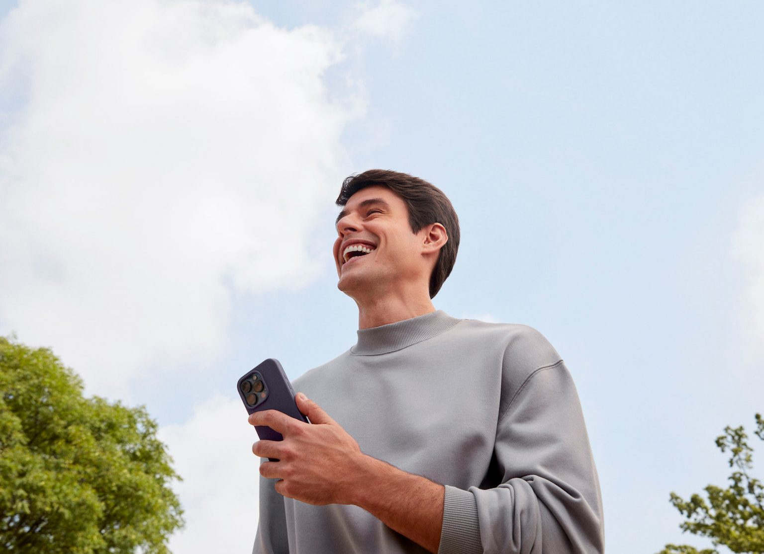 Homem branco sorrindo olhando para frente enquanto segura com a mão esquerda um celular, em um ambiente externo a luz do dia com nuvens no céu e uma árvore a sua esquerda e outra árvore a sua direita.