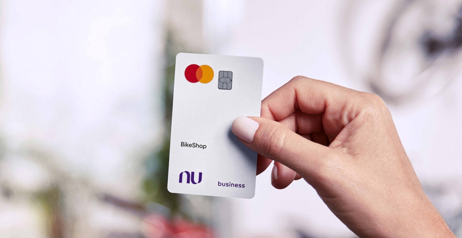 Mão direita de uma pessoa, com pulseira de pérolas no pulso, segura um cartão Nubank business branco. Ao fundo estão várias bicicletas de cores variadas desfocadas.