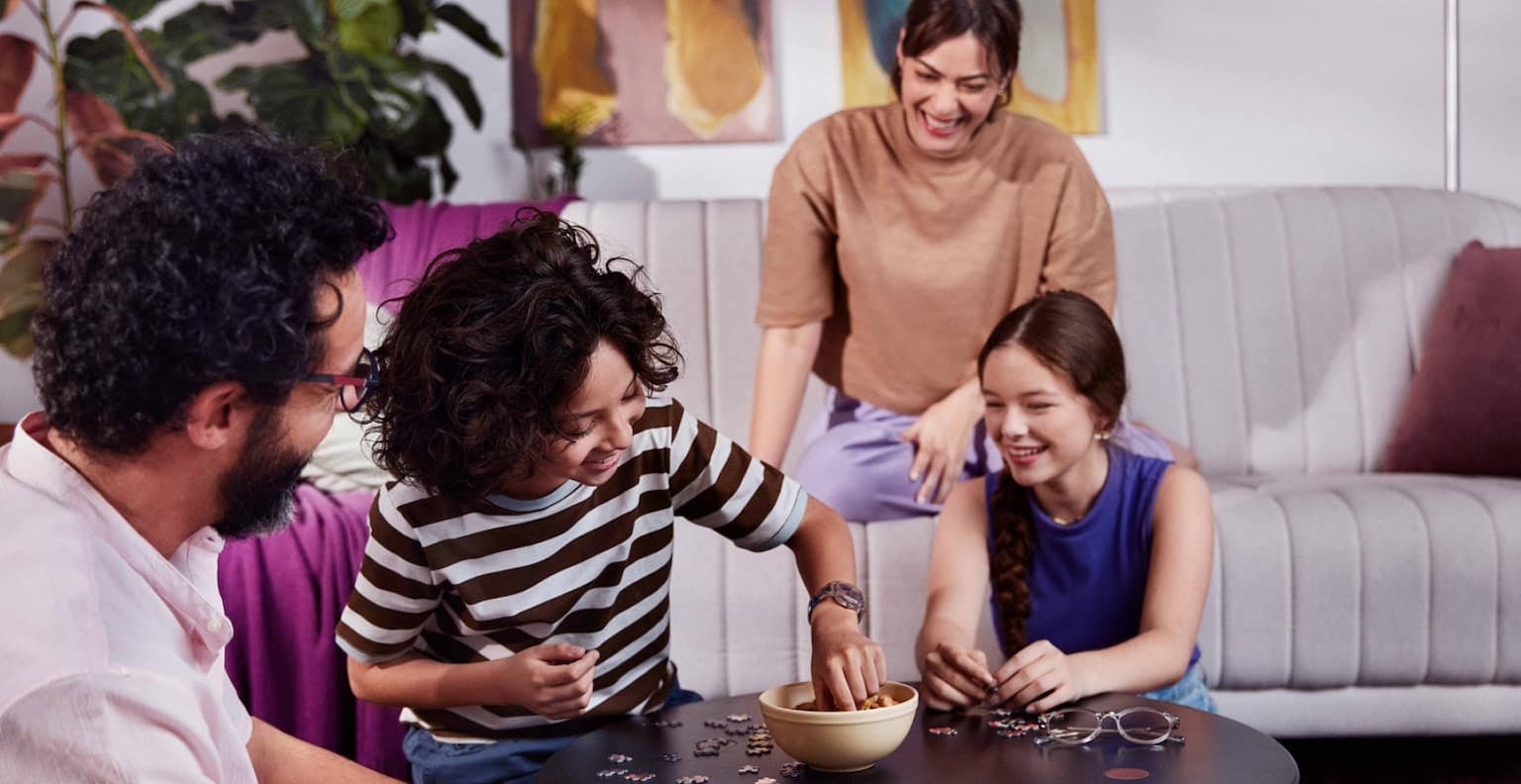 Quatro pessoas em uma sala, homem sentado no chão olha para duas crianças sorrindo e sentadas no chão em volta de uma mesa de centro enquanto brincam com peças de um quebra-cabeça, ao fundo uma mulher sentada no sofá observa as crianças enquanto sorri.