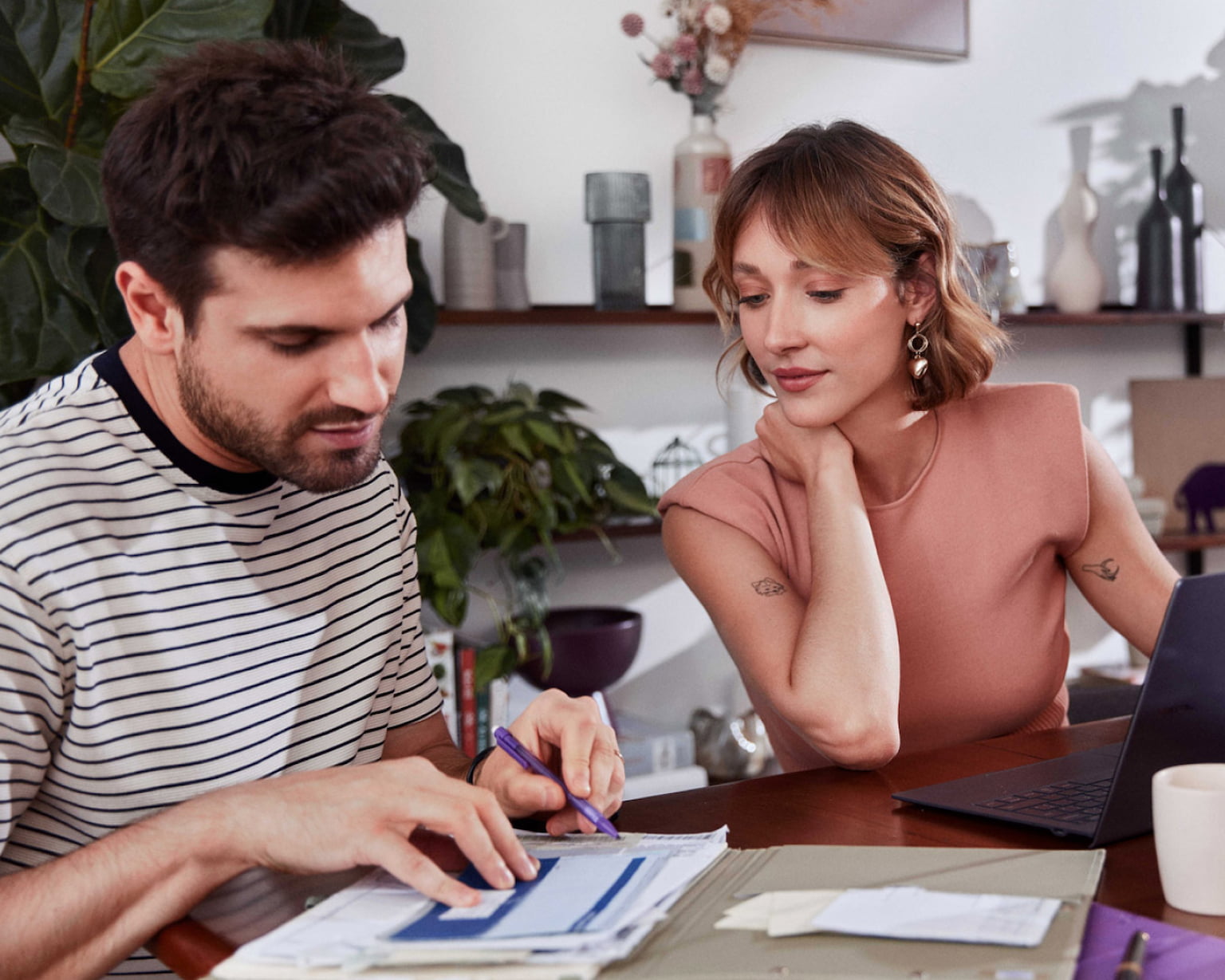 Sentado em uma mesa de madeira um homem branco está lendo alguns papéis enquanto aponta com a caneta para o documento, ao seu lado esquerdo uma mulher branca com a mão esquerda em cima de um caderno observa o que ele está mostrado, a sua frente um notebook, um copo com liquido roxo dentro e uma xícara pequena.
