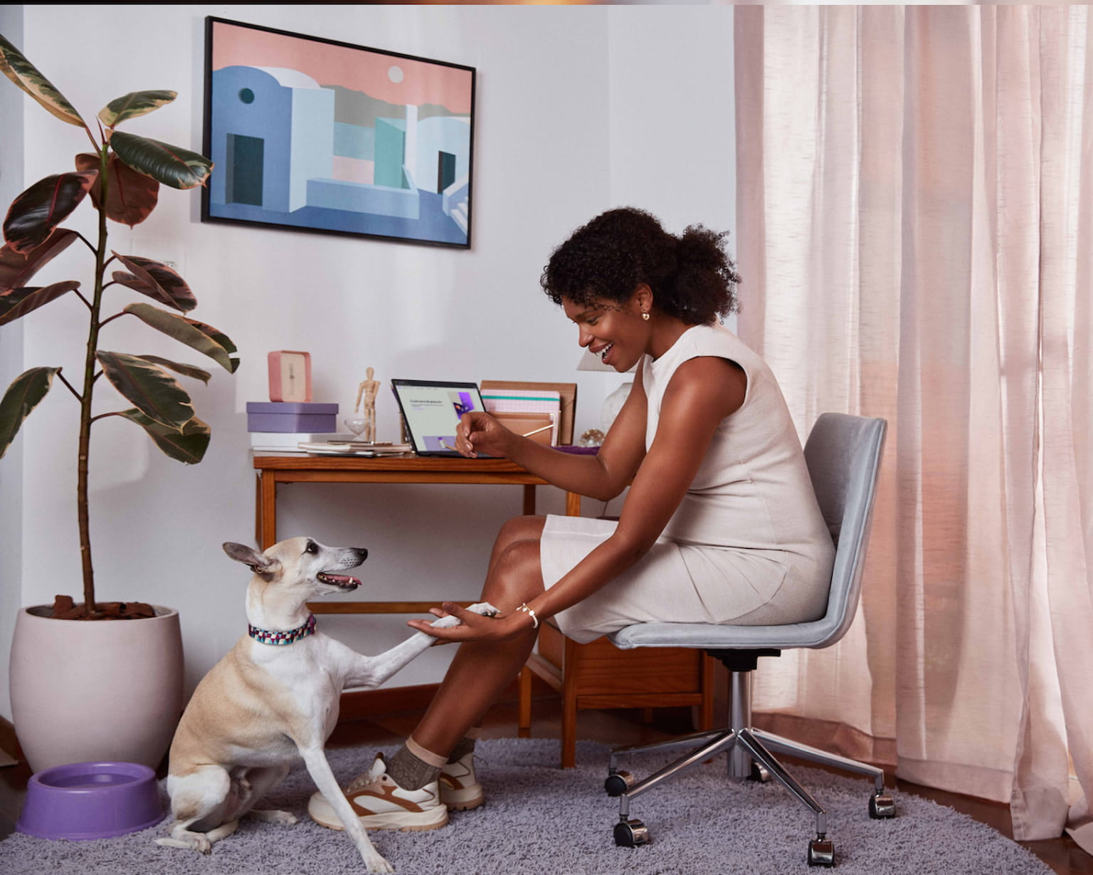Mulher negra sentada em uma cadeira de escritório, sorrindo, segurando com a mão direita a pata esquerda de um pequeno cachorro que está sentado no chão próximo de um vaso grande de plantas e uma tigela de água, há uma cortina na parede direita, ao fundo uma mesa com um notebook e um quadro na parede com um desenho de casas.