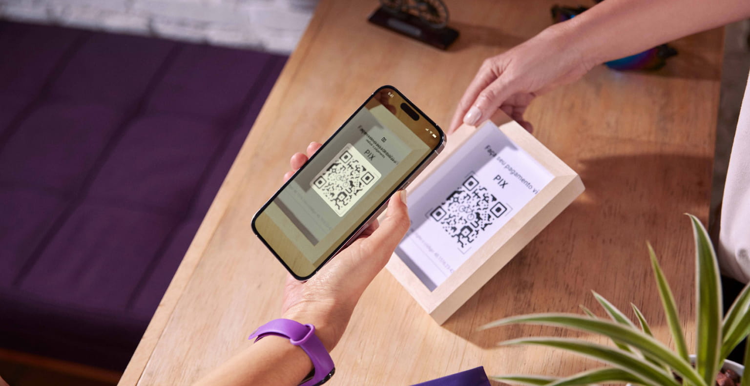 Pessoa com relógio roxo lendo QR code para pagamento por pix com seu celular. Do outro lado, uma mão segura um quadro pequeno com o código para o pagamento.