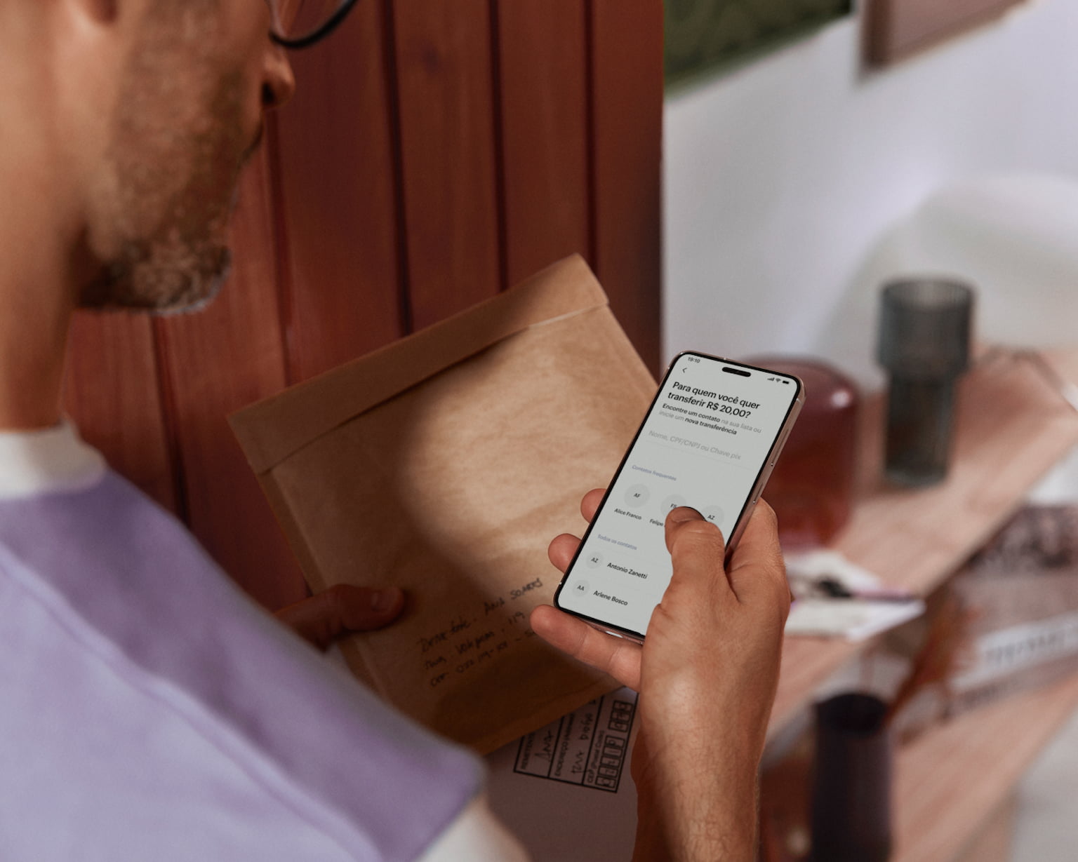 Parte das costas de um homem branco segurando o celular e visualizando sua conta no aplicativo do Nubank com a mão direita e com a esquerda segura um envelope e uma caixa branca.