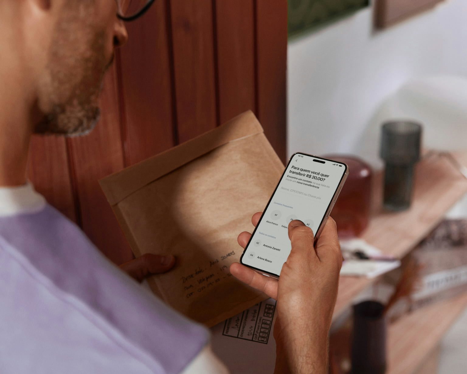 Parte das costas de um homem branco segurando o celular e visualizando sua conta no aplicativo do Nubank com a mão direita e com a esquerda segura um envelope e uma caixa branca.