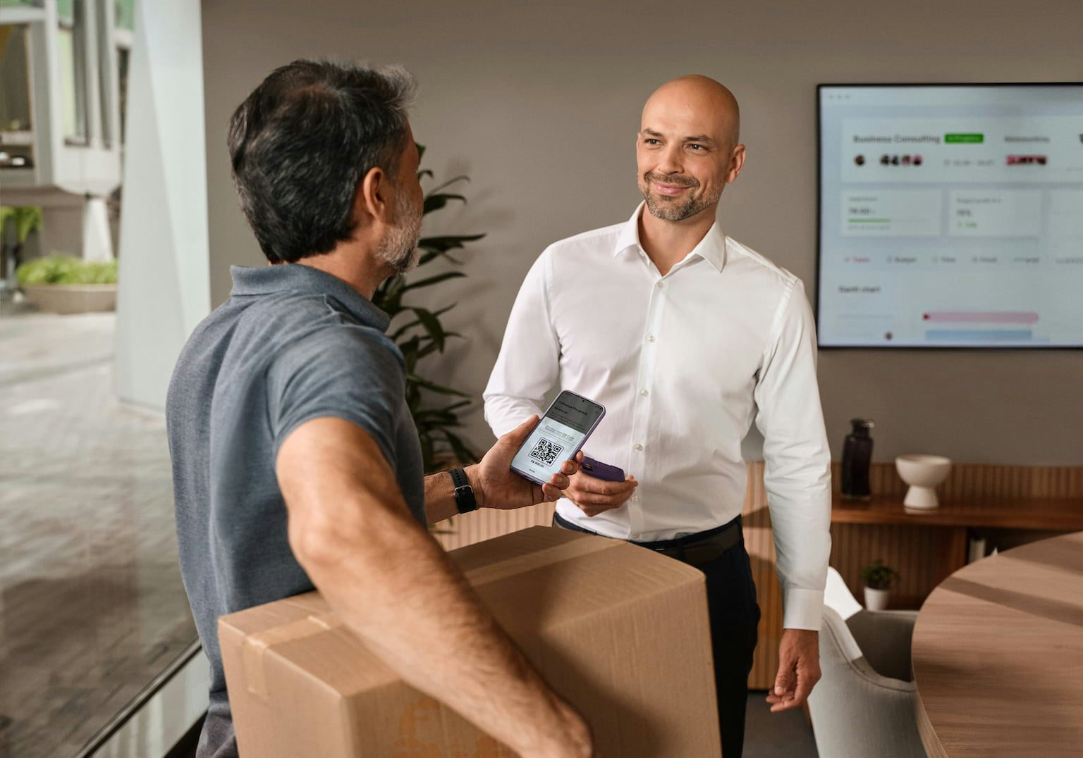 Em primeiro plano Imagem mostra um homem de costas vestindo uma camisa cinza segurando uma caixa de papelão em um dos braços e um celular na outra mão. Na tela é possível ver um QRCode. Ele olha para outro homem, que está de frente, sorrindo, e segurando um celular em uma das mãos. Ele veste uma camisa branca e calça preta.