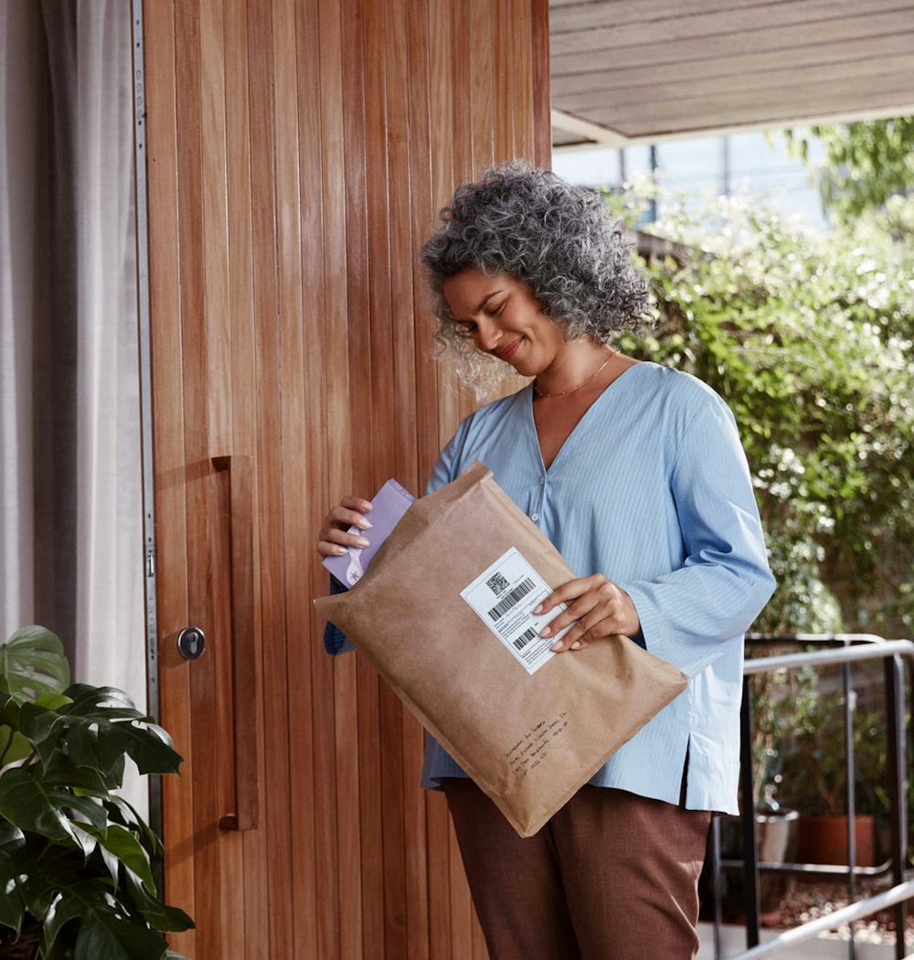Mulher negra de cabelos cacheados e grisalhos, em pé do lado de uma porta de madeira, segurando um envelope e sorrindo enquanto retira um livro de dentro, a sua frente há uma parede branca com um quadro abstrato, interruptores e uma luz de parede, no chão algumas folhagens de uma planta em um vaso.
