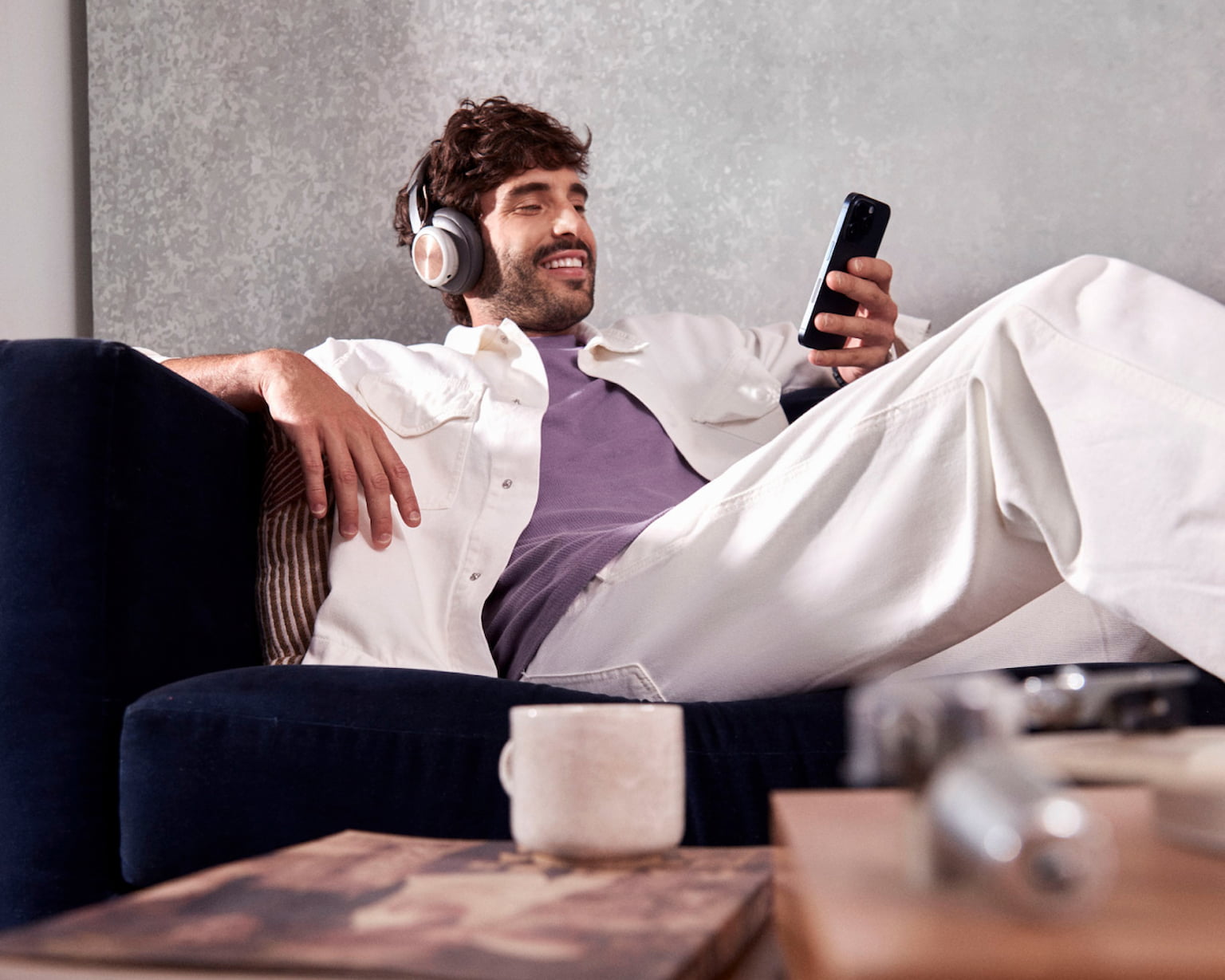 Em um plano desfocado uma mesa de madeira com uma pequena xícara de café em cima de um livro, uma caixa de madeira ao lado e um cartão do Nubank, ao fundo em um plano nítido um homem branco sentado confortavelmente em um sofá, sorrindo enquanto usa um headset e olha para o celular em uma de suas mãos.