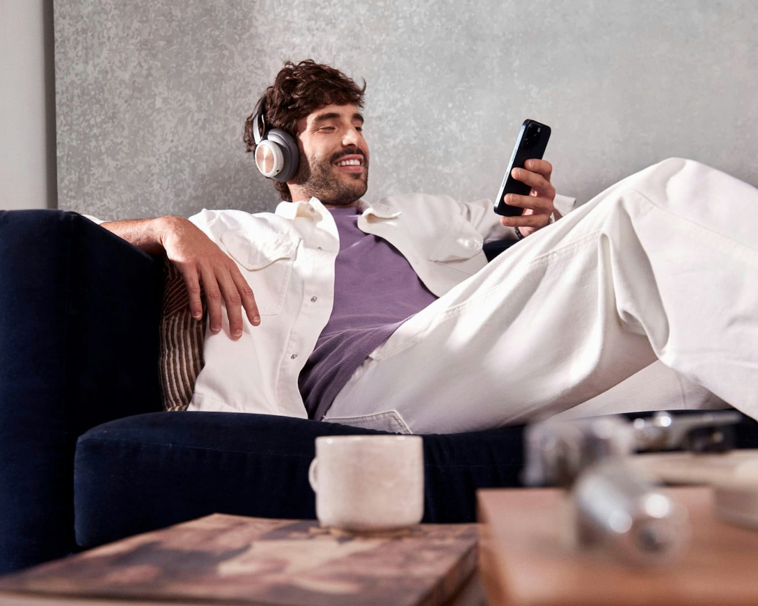 Em um plano desfocado uma mesa de madeira com uma pequena xícara de café em cima de um livro, uma caixa de madeira ao lado e um cartão do Nubank, ao fundo em um plano nítido um homem branco sentado confortavelmente em um sofá, sorrindo enquanto usa um headset e olha para o celular em uma de suas mãos.