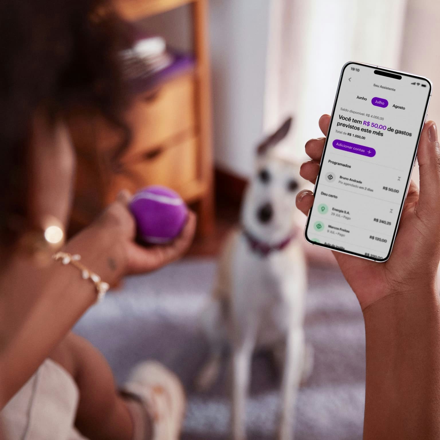 Vista das costas de uma mulher negra sentada de pernas cruzadas, com um celular na mão direita e visualizando sua conta no aplicativo do Nubank, com sua mão esquerda segura uma bolinha de tênis roxa, ao fundo e desfocado um cachorro de pequeno porte sentado no chão olhando para a bolinha.