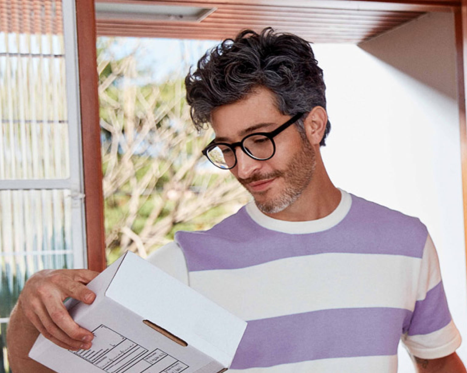 Homem branco usando óculos, entrando em um ambiente fechado por uma porta de madeira enquanto olha para uma caixa branca que está na sua mão direita, sua mão esquerda está na maçaneta da porta e segura um envelope.