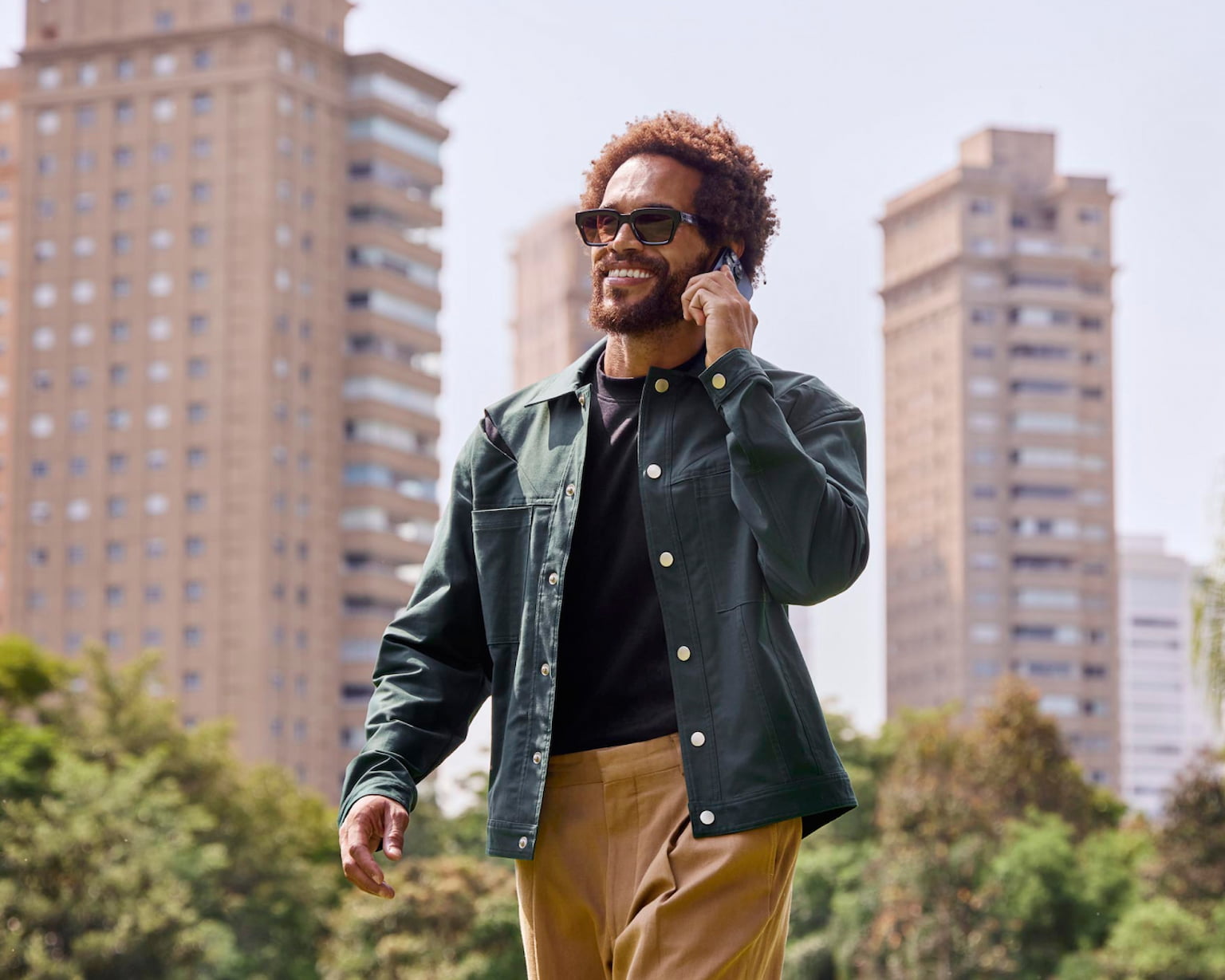 Homem sorrindo enquanto fala no celular passeando pela cidade. Ele está segurando o celular com a mão esquerda e, ao fundo, prédios e algumas árvores.