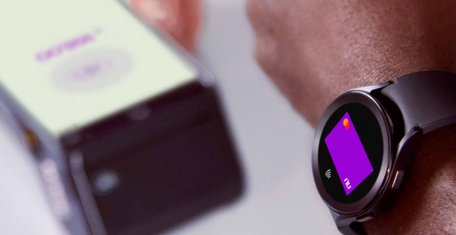 Braço de uma pessoa negra usando um smartwatch para efetuar o pagamento com seu cartão virtual Nubank por aproximação, do outro lado a mão de uma pessoa branca segurando uma máquina de cartão de crédito em cima de uma mesa com um óculos e um capacete de ciclista em cada lado.