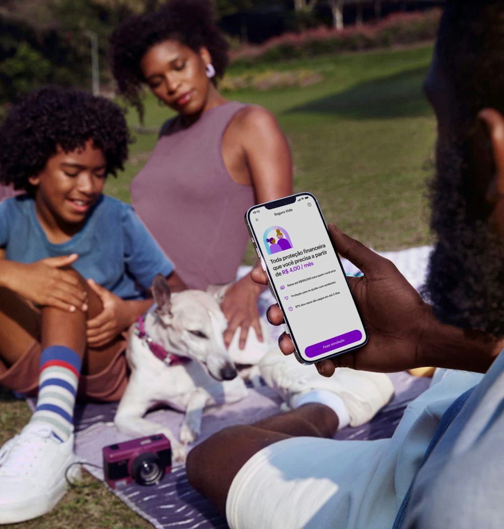 Homem sentado segurando um celular com o aplicativo do Nubank aberto. Ao fundo está sua família e seu cachorro. Todos estão sentados no chão, em cima de um pano com detalhes roxos, onde também há uma câmera fotográfica roxa.