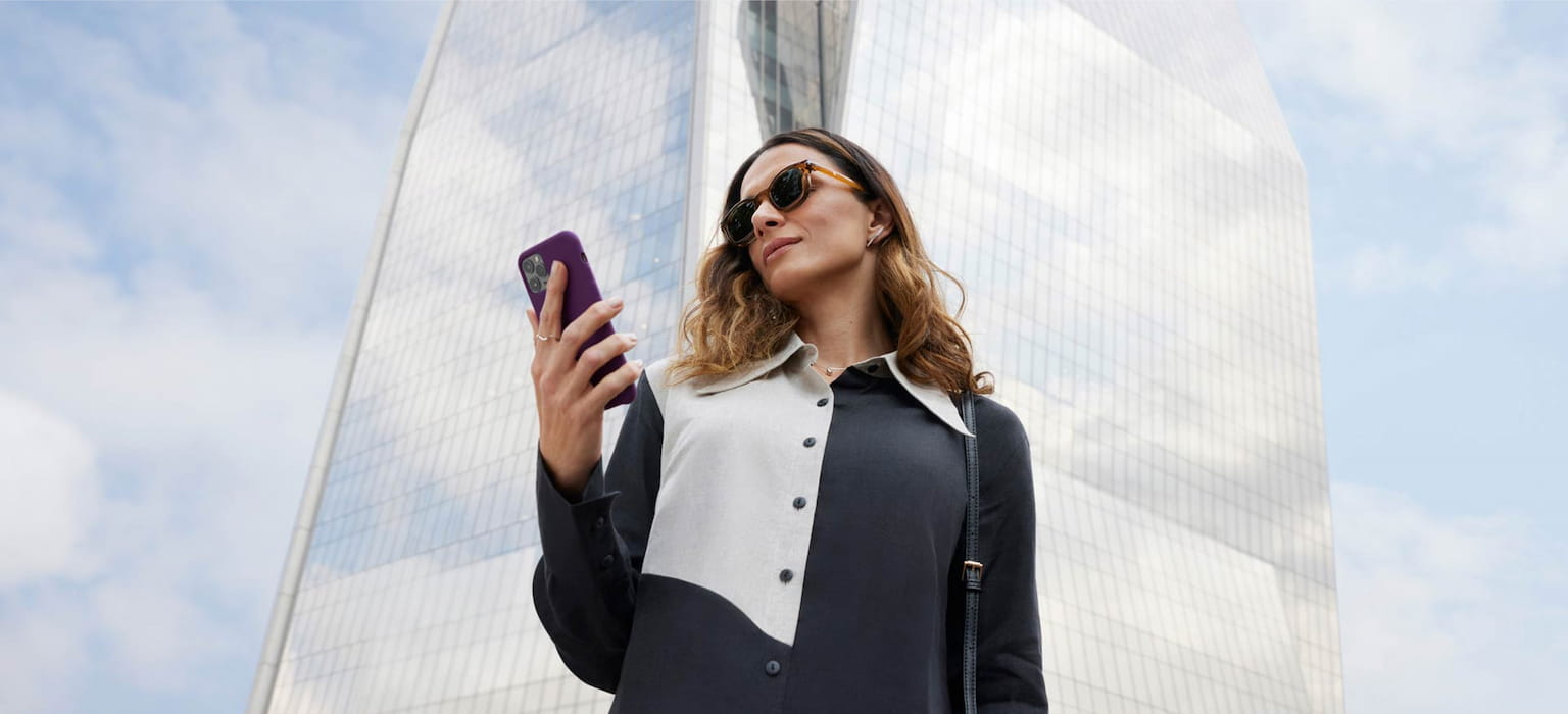 Mulher de óculos escuro, camisa preta com detalhes em branco olhando para o celular. Ao fundo, um prédio arquitetônico coberto por vidros que espelham as nuvens e o azul do céu.