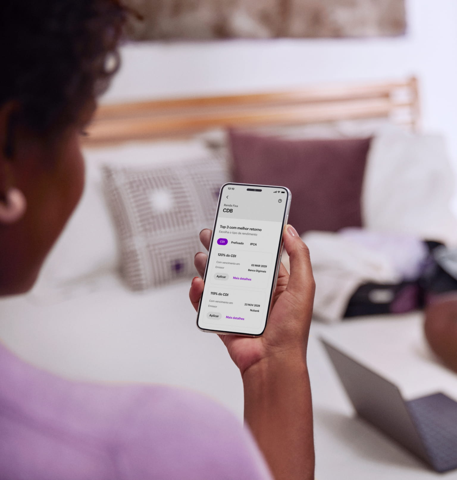 Vista das costas de uma mulher negra sentada, com um celular na mão direita e visualizando sua conta no aplicativo do Nubank, ao fundo e desfocado um computador em cima da cama