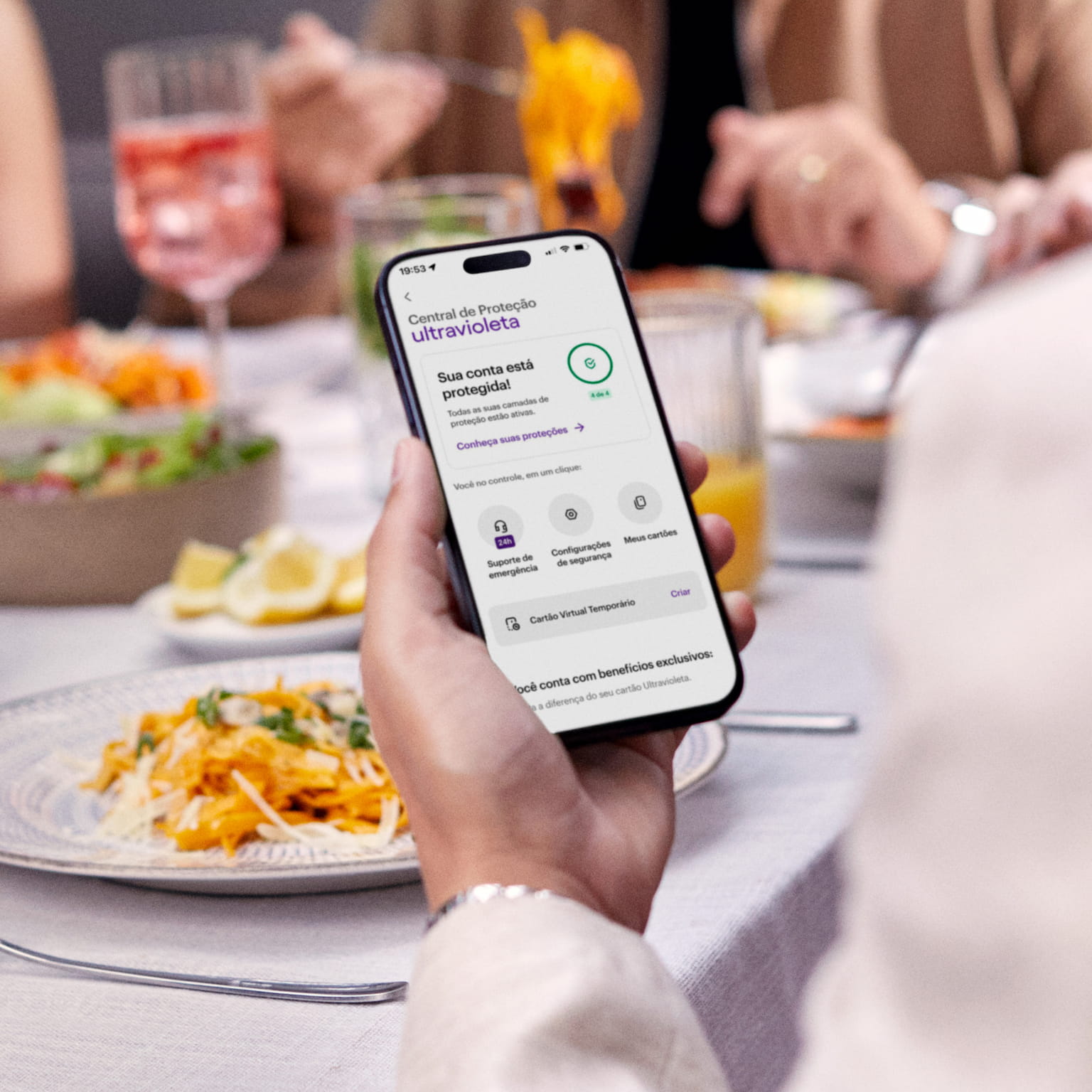 Homem de costas olha para o celular, que está em sua mão esquerda sobre uma mesa com drinks e comidas. Ele está checando a Central de Proteção Ultravioleta, no App do Nu. Ao fundo e de frente para ele, um casal rindo enquanto almoçam.
