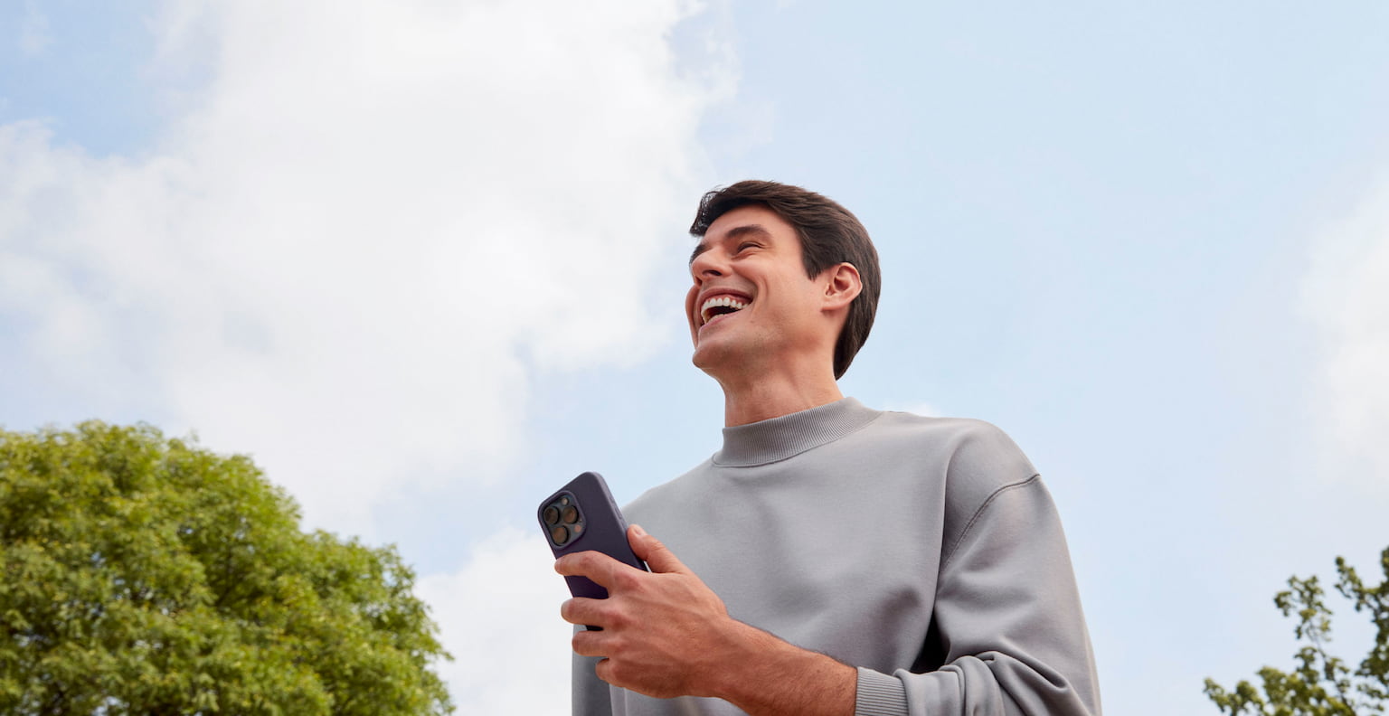 Homem branco sorrindo olhando para frente enquanto segura com a mão esquerda um celular, em um ambiente externo a luz do dia com nuvens no céu e uma árvore a sua esquerda e outra árvore a sua direita.