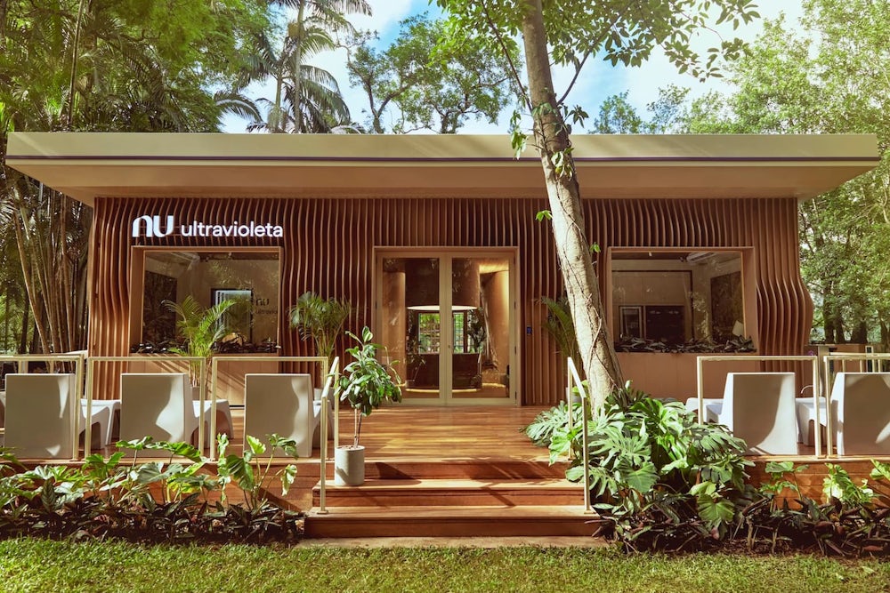 Foto da fachada da Casa Nubank Ultravioleta, no Ibirapuera. Toda de madeira, com o logo do Nu e o nome Ultravioleta na frente. Com uma pequena área verde na frente.