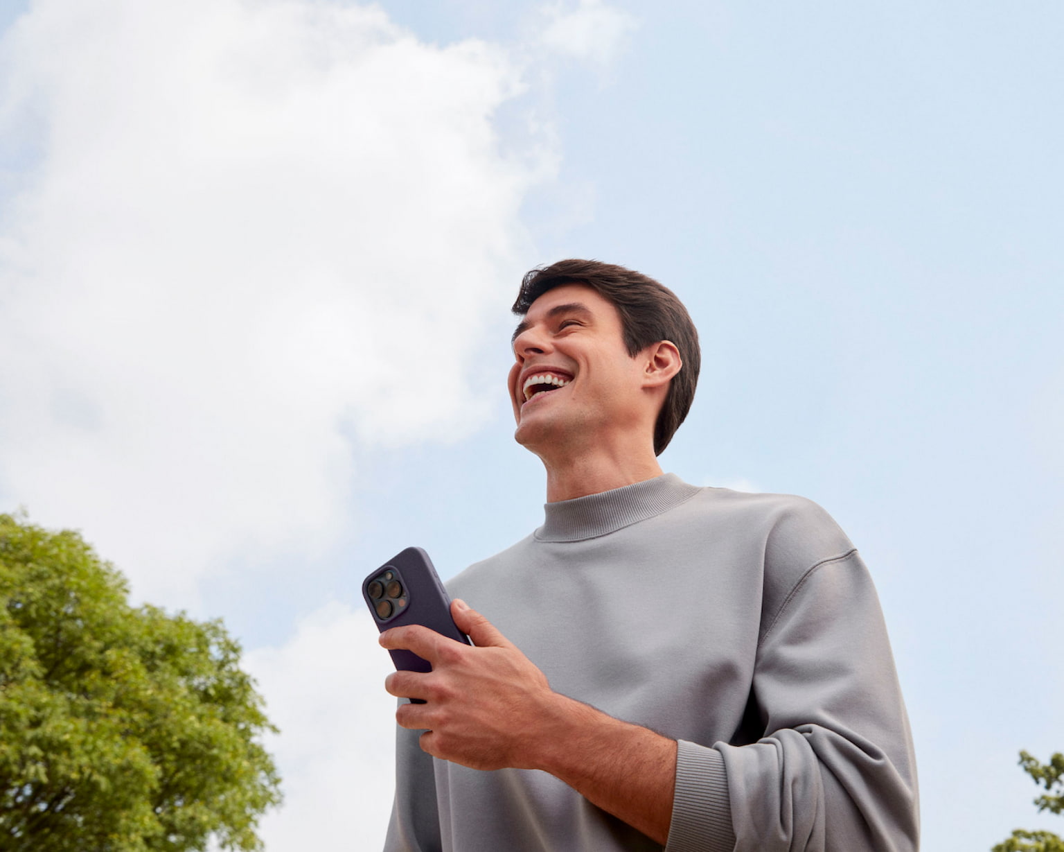Homem branco sorrindo olhando para frente enquanto segura com a mão esquerda um celular, em um ambiente externo a luz do dia com nuvens no céu e uma árvore a sua esquerda e outra árvore a sua direita.