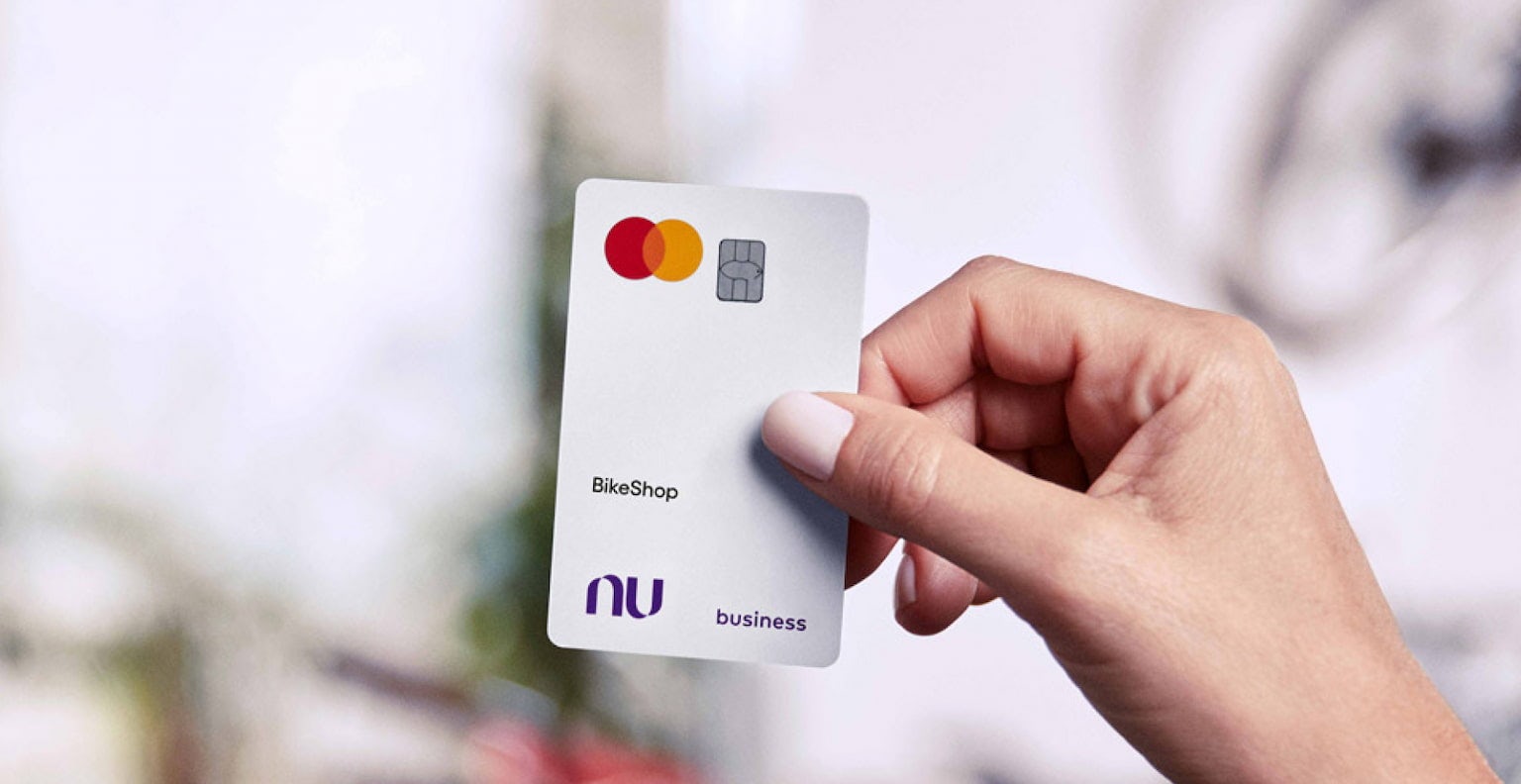 Mão direita de uma pessoa, com pulseira de pérolas no pulso, segura um cartão Nubank business branco. Ao fundo estão várias bicicletas de cores variadas desfocadas.