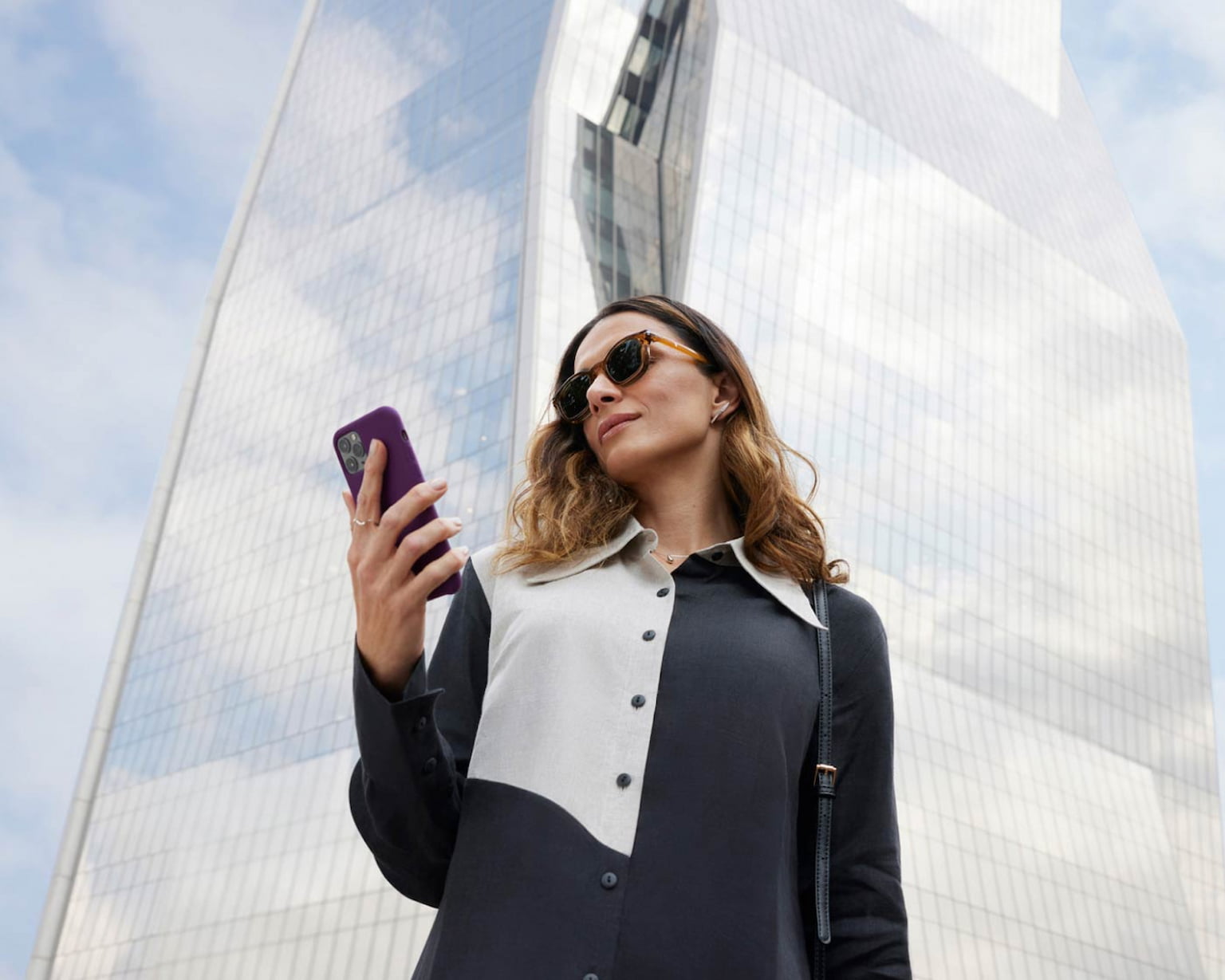 Mulher de óculos escuro, camisa preta com detalhes em branco olhando para o celular. Ao fundo, um prédio arquitetônico coberto por vidros que espelham as nuvens e o azul do céu.
