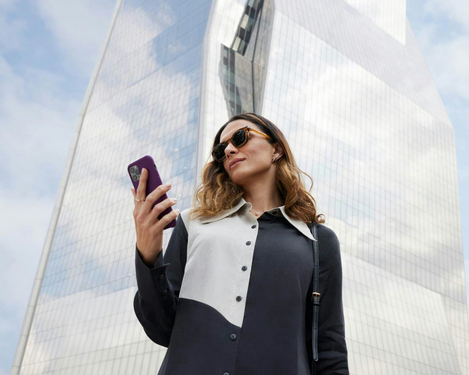 Mulher de óculos escuro, camisa preta com detalhes em branco olhando para o celular. Ao fundo, um prédio arquitetônico coberto por vidros que espelham as nuvens e o azul do céu.