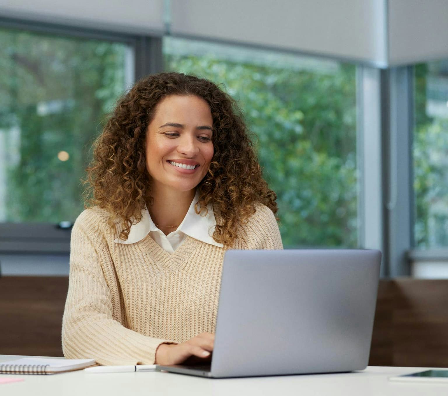 Mulher de cabelos cacheados sorrindo enquanto digita no notebook que está em cima de uma mesa com alguns objetos de trabalho. Ao fundo, janelas para uma área externa com plantas.