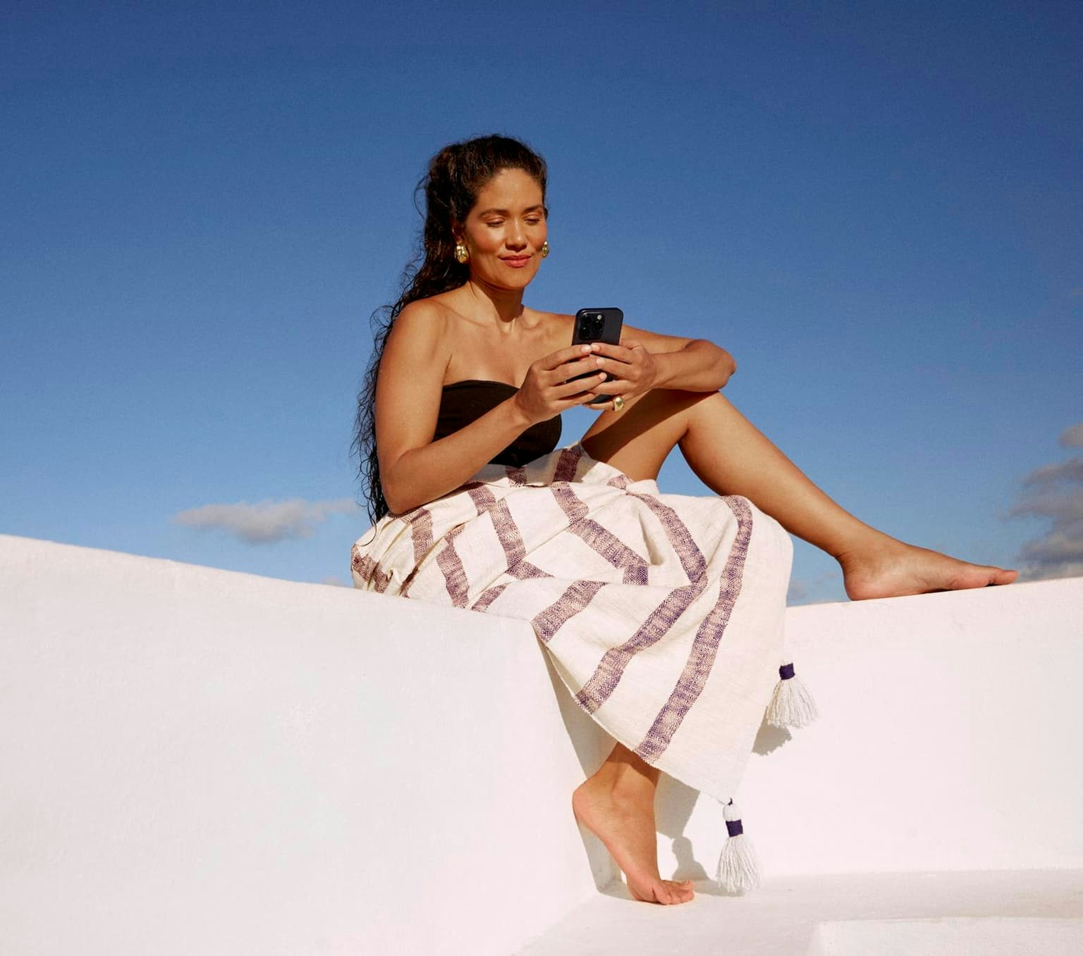 Mulher de top e uma canga sobre as pernas está sentada em uma superfície branca na luz do dia com o céu azul. Ela segura o celular com as duas mãos e olha para a tela com um sorriso leve.