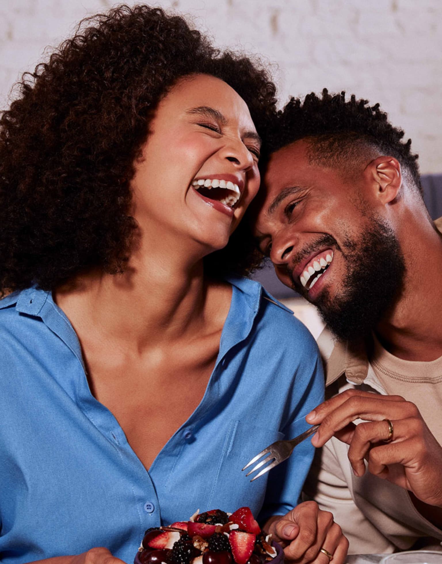 Uma mulher negra à esquerda ri segurando uma tigela com frutas. À sua direita um homem negro ri encostado nela com um garfo em direção à sua tiigela. Ambos estão sentados à uma mesa de café da manhã.