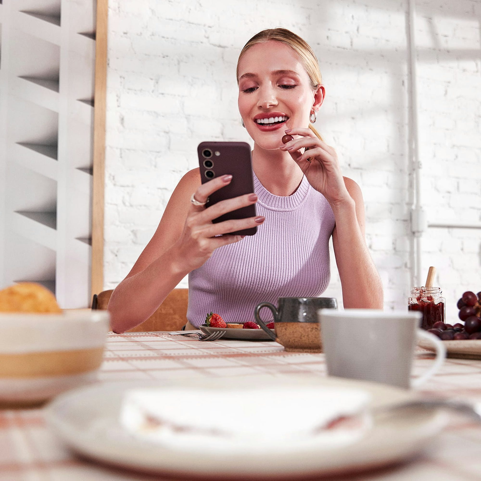 Mulher branca com o cabelo preso está sentada em frente a uma mesa com um prato com tapioca, xícaras, morangos, uvas e geleia. Ela olha para o celular em uma das mãos, enquanto leva uma uva a sua boca com a outra mão. Ao fundo, uma parede de tijolos brancos com um quadro abstrato.