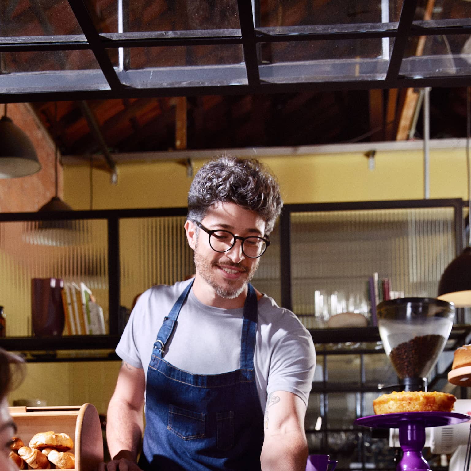 Um homem de barba e cabelo grisalho, com óculos redondo de grau, usando uma camisa cinza e um avental jeans está olhando para baixo. Do seu lado direito há um suporte de madeira com croissants, e do lado esquerdo, um bolo e uma cafeteira com grãos de café. Ao fundo, há uma prateleira com taças e livros.