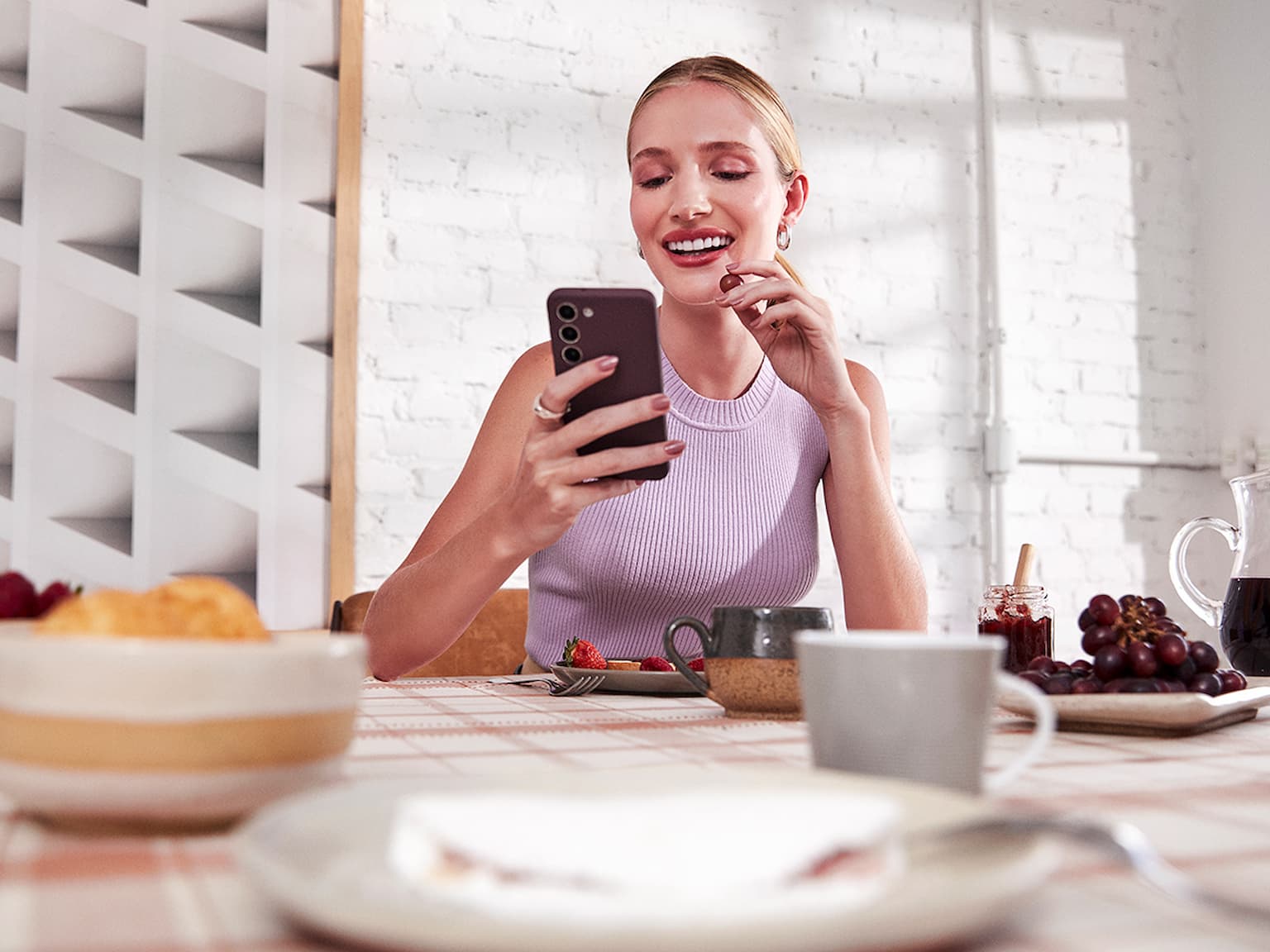 Mulher branca com o cabelo preso, sentada em frente a uma mesa com alguns pratos, xícaras, tigelas com frutas e uma jarra de suco. Ela olha para o celular, que está em sua mão direita, enquanto segura uma uva com a mão esquerda. Ao fundo, uma parede de tijolos brancos e um quadro abstrato, ambos iluminados pela luz do dia.