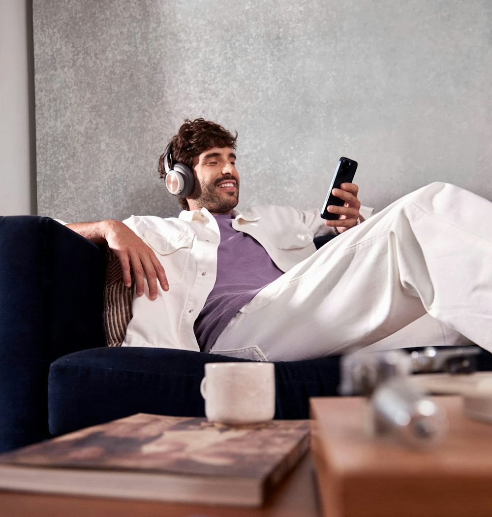 Homem branco sentado confortavelmente em um sofá, sorrindo enquanto usa um headset e olha para o celular em uma de suas mãos