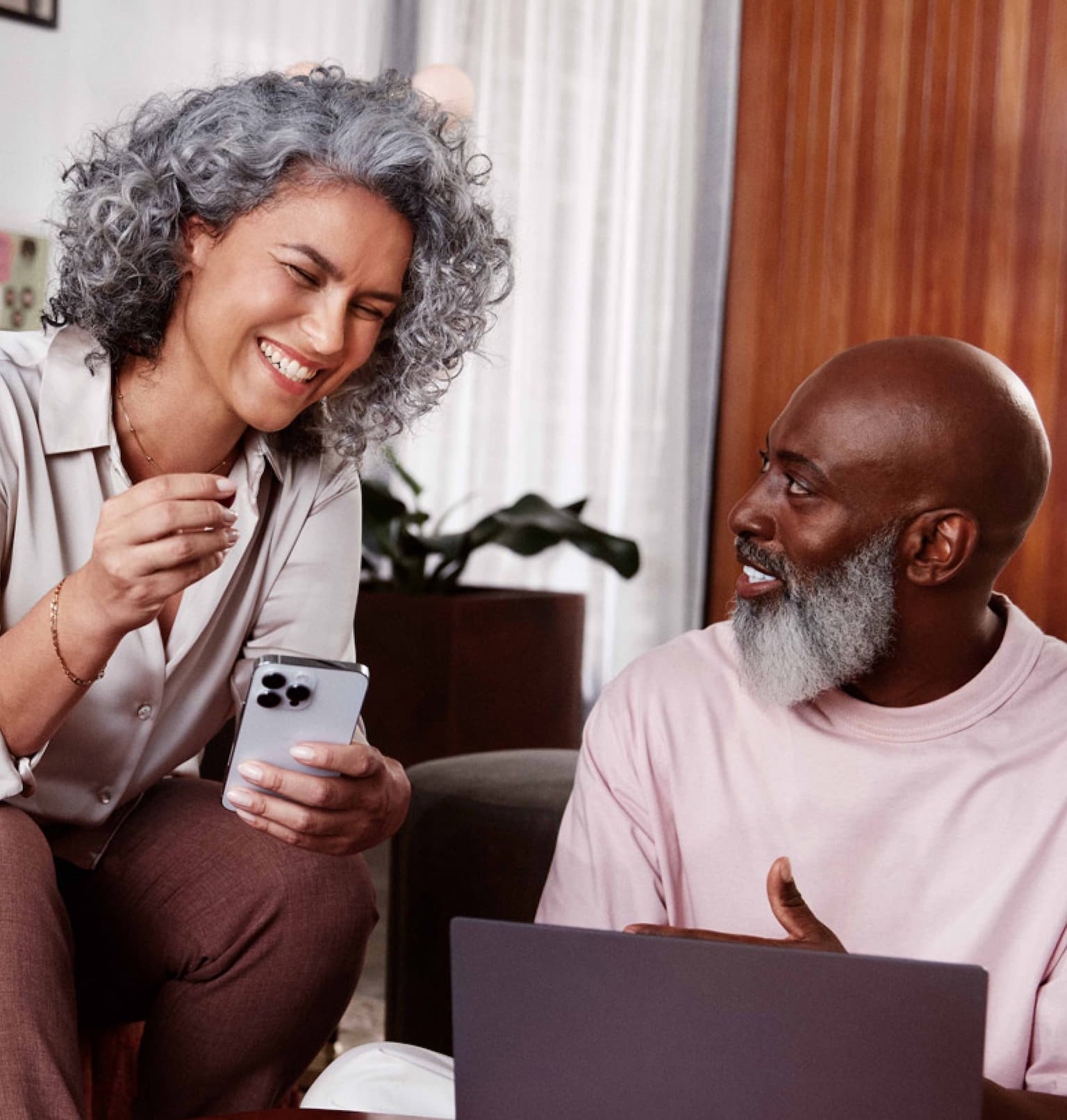 Mulher negra de cabelos cacheados e grisalhos sentada em um sofá e sorrindo, segurando com a mão esquerda um celular em direção a um homem negro, careca, com barba grisalha, sentado ao seu lado no chão, olhando para o rosto dela enquanto sorri e com uma pequena mesa de madeira a sua frente com um notebook.