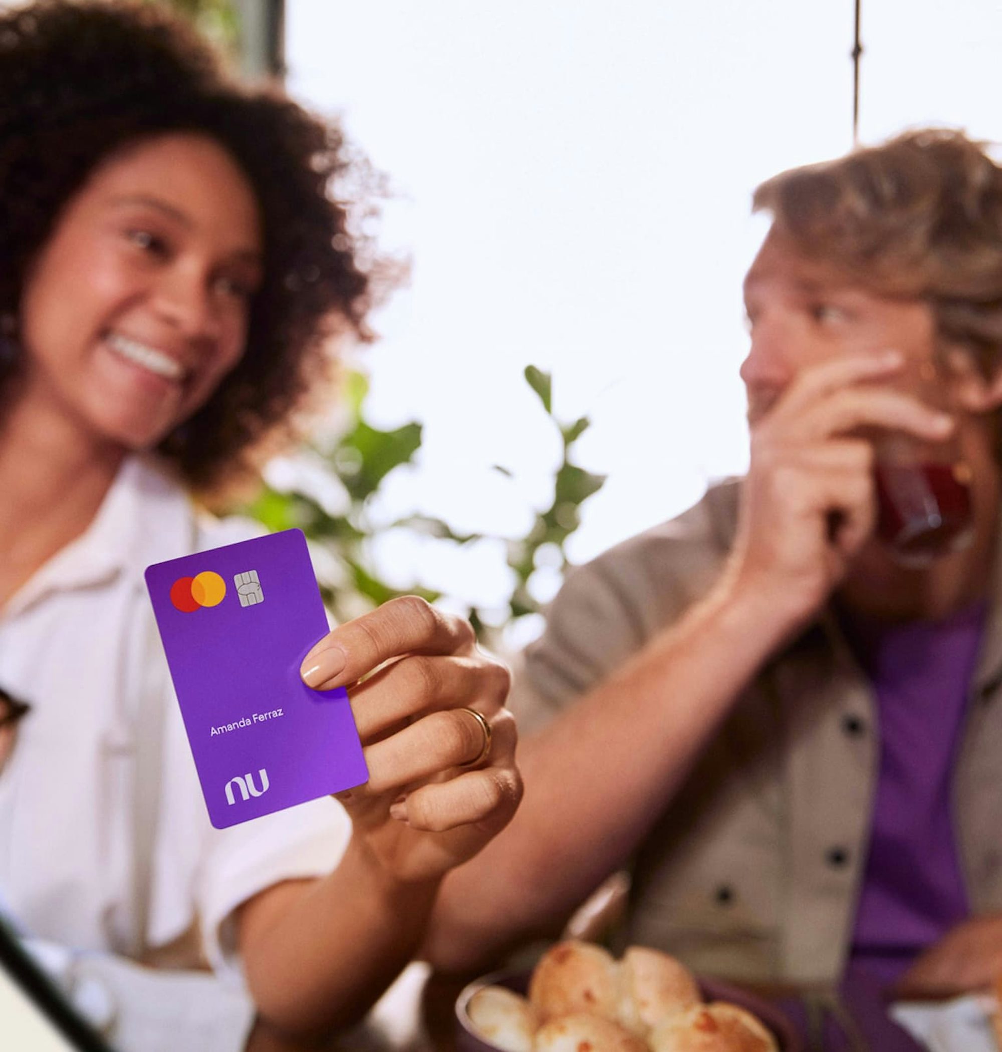 Mulher negra mostrando um cartão Nubank na direção do visualizador da imagem. À seu lado há um homem branco. Ambos estão sentados à mesa com pães de queijo. Toda a imagem é desfocada com exceção do cartão Nubank.