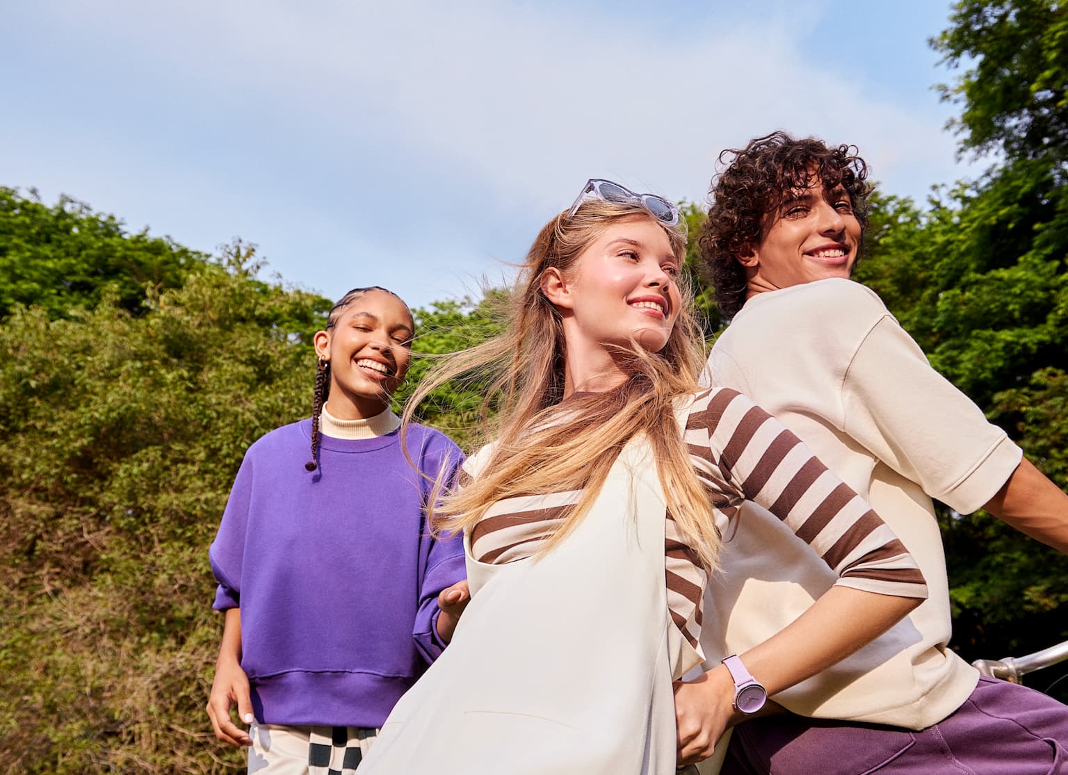 Duas meninas e um menino sorrindo em um parque. Uma das meninas está vestindo uma blusa roxa e a outra está com uma blusa bege com listras marrons horizontais com um vestido bege por cima. O menino está de costas, em cima de uma bicicleta, vestindo uma camiseta bege e uma calça roxa.