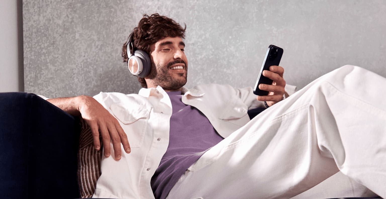 Homem branco sentado confortavelmente em um sofá, sorrindo enquanto usa um headset e olha para o celular em uma de suas mãos