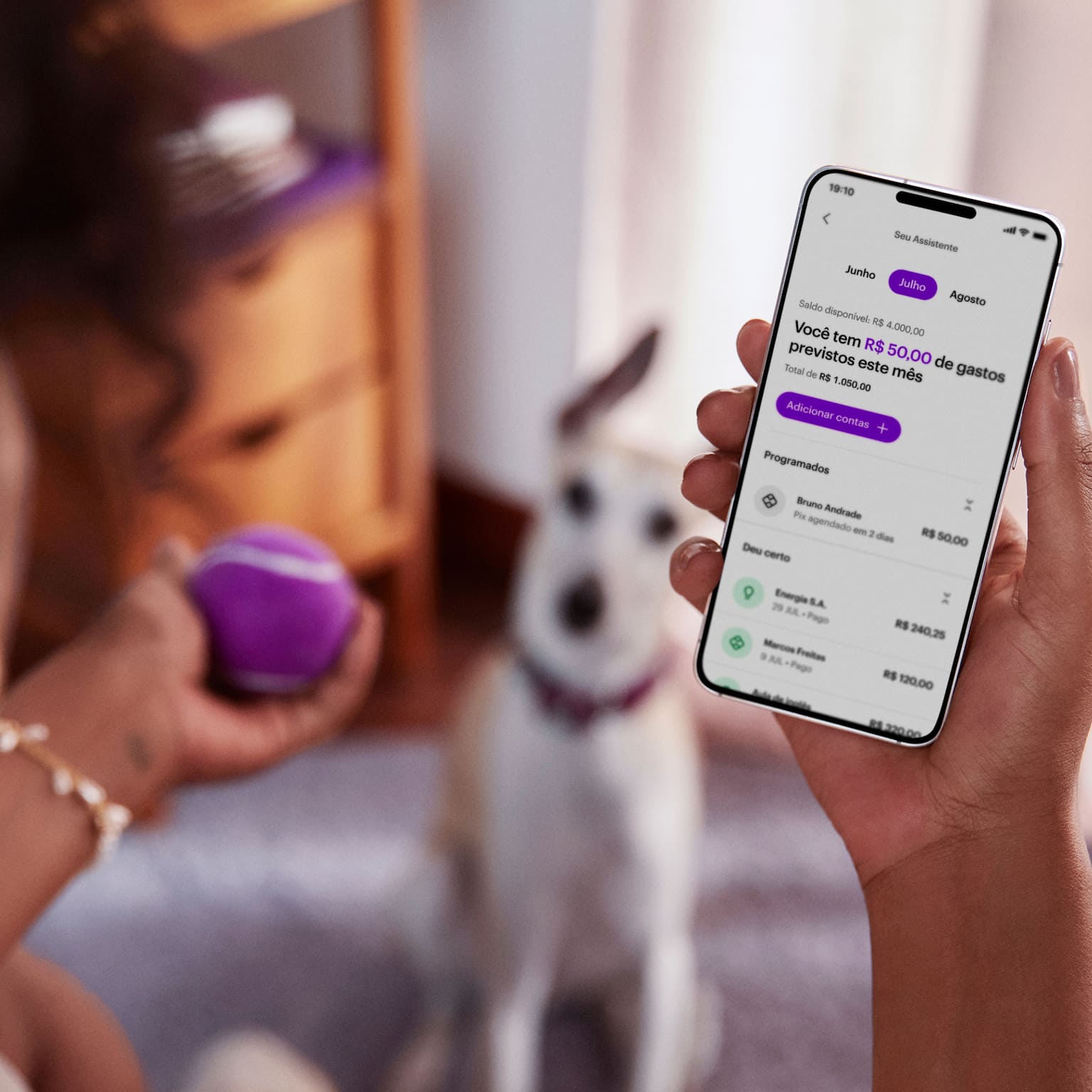 Vista das costas de uma mulher negra sentada de pernas cruzadas, com um celular na mão direita e visualizando sua conta no aplicativo do Nubank, com sua mão esquerda segura uma bolinha de tênis roxa, ao fundo e desfocado um cachorro de pequeno porte sentado no chão olhando para a bolinha.