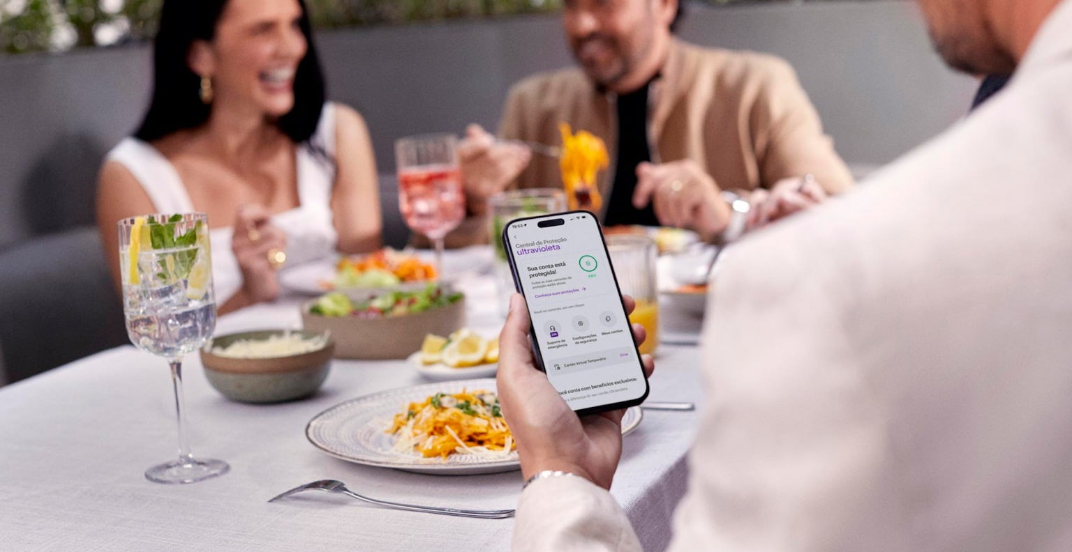 Homem de costas olha para o celular, que está em sua mão esquerda sobre uma mesa com drinks e comidas. Ele está checando a Central de Proteção Ultravioleta, no App do Nu. Ao fundo e de frente para ele, um casal rindo enquanto almoçam.
